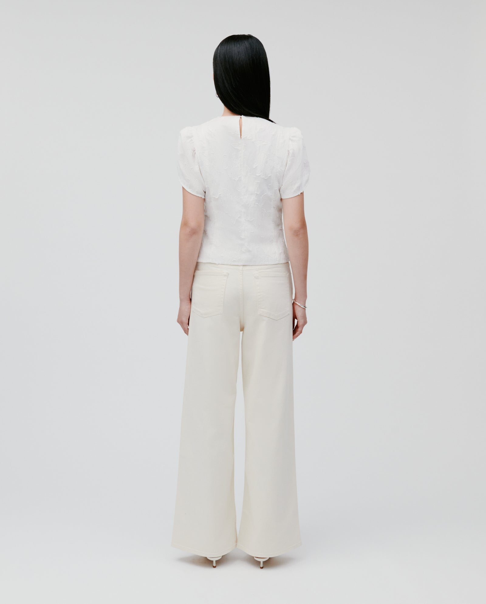 A woman stands facing away in a plain studio, wearing the IVY OAK ELODIE Blouse with voluminous sleeves and wide-leg cream pants.