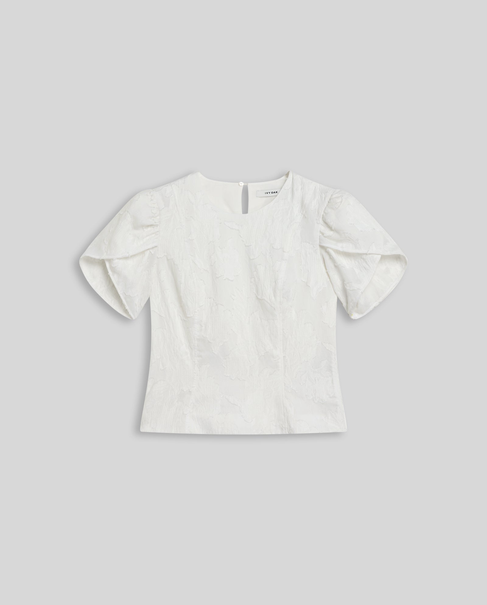 The ELODIE Blouse by IVY OAK features a white fil coupé fabric with a subtle floral texture and voluminous puffed sleeves, creating a modern romantic silhouette.
