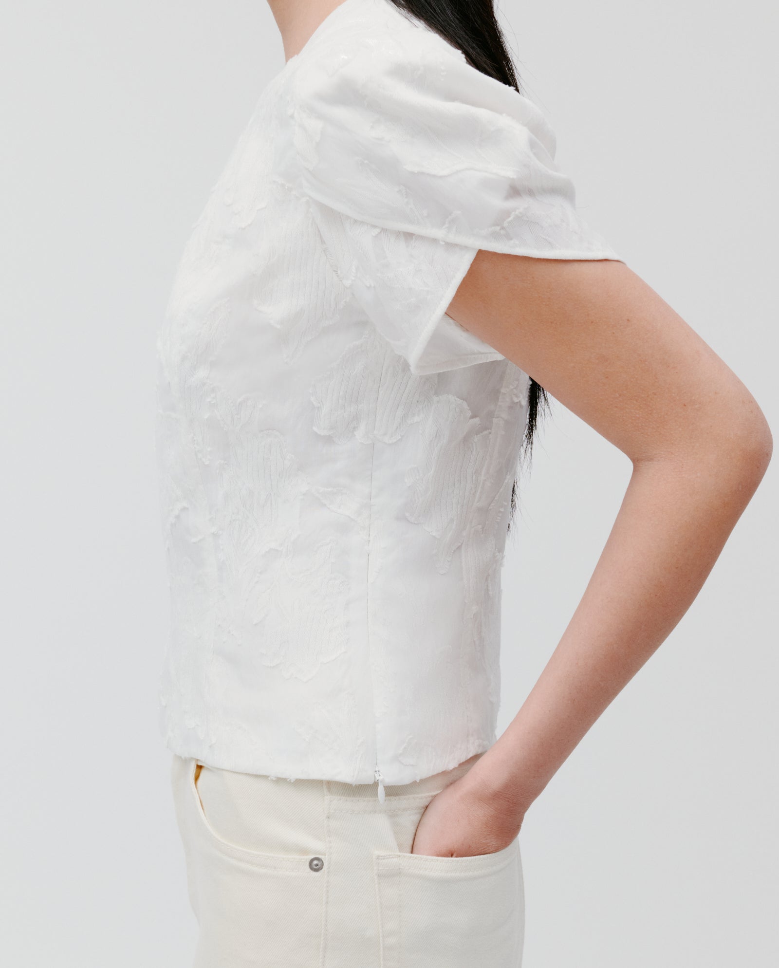 A person wearing the IVY OAK ELODIE Blouse, featuring textured white fil coupé fabric and voluminous sleeves, paired with white pants. Hand in pocket, side view.