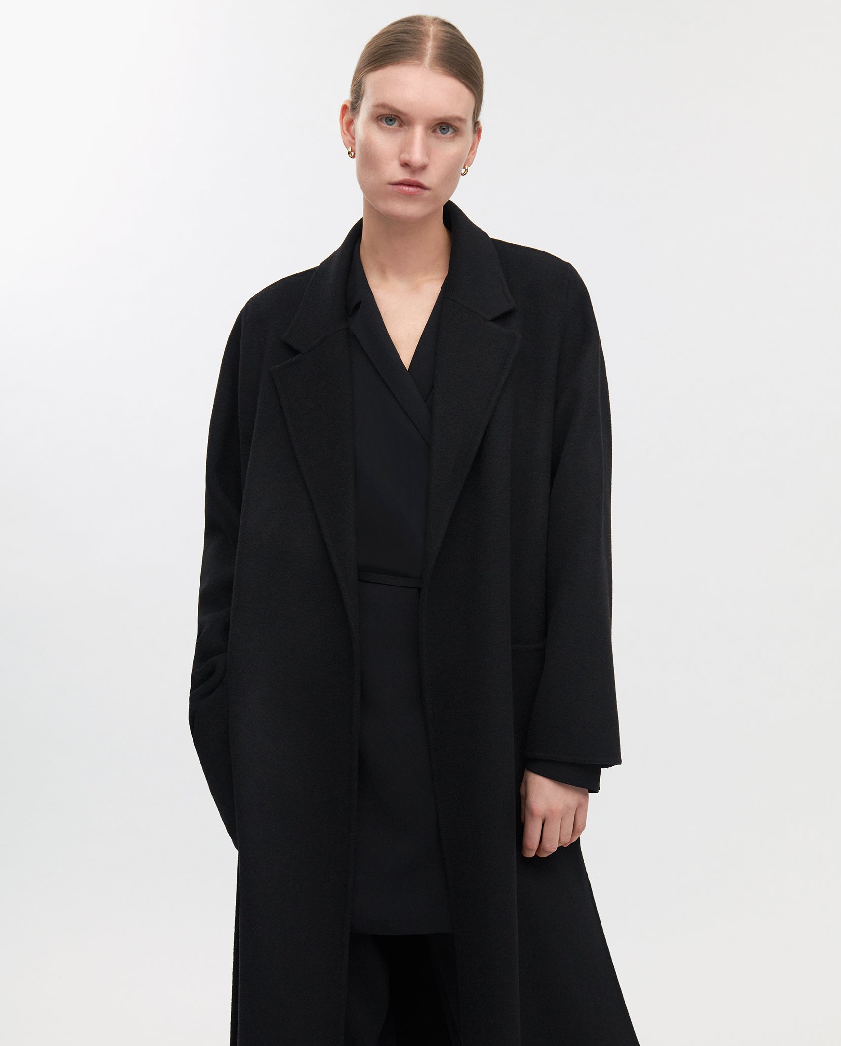 A person stands against a plain white background wearing the IVY OAK CELIA Doubleface Coat, a long black midi-length wool coat crafted to Responsible Wool Standard, paired with a matching black outfit.