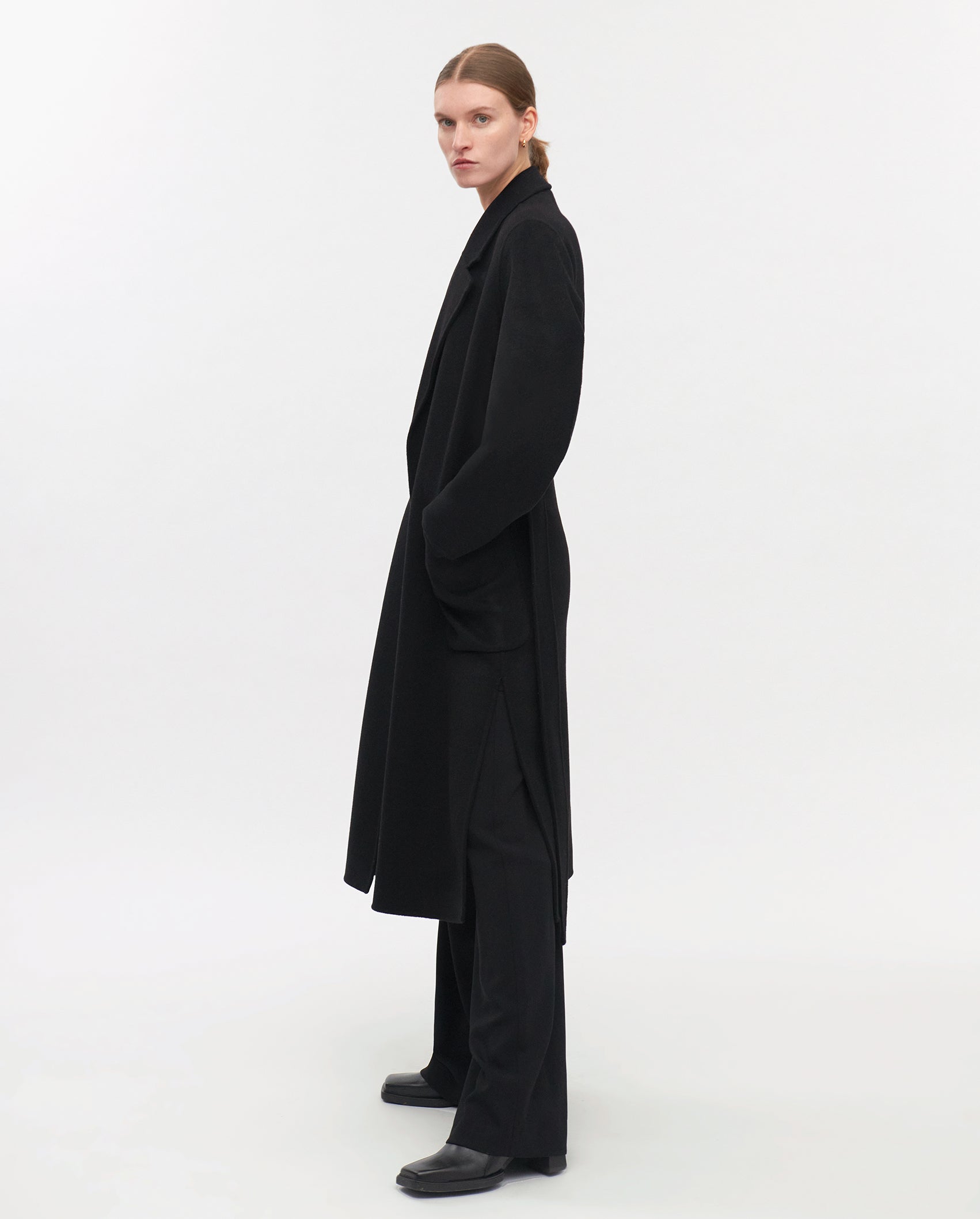 Wearing the IVY OAK CELIA Doubleface Coat, a black midi-length wool coat certified by the Responsible Wool Standard, styled with black pants and shoes, standing against a plain white background.