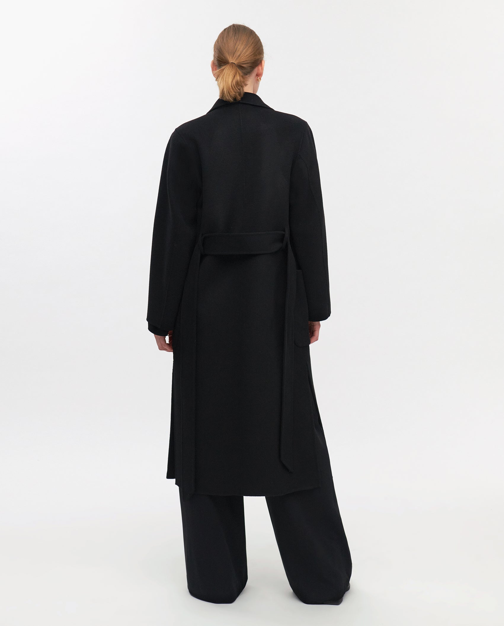 Person wearing the IVY OAK CELIA Doubleface Coat, a long black wool midi coat made to the Responsible Wool Standard, paired with wide black pants. Shown from the back on a white background.