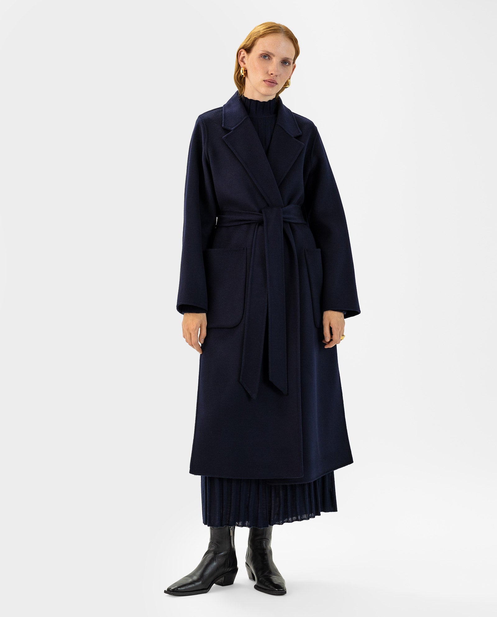A woman wears the IVY OAK CELIA Doubleface Coat in navy, made to Responsible Wool Standard guidelines, with a belt over a navy pleated midi dress and black boots, standing against a white background.