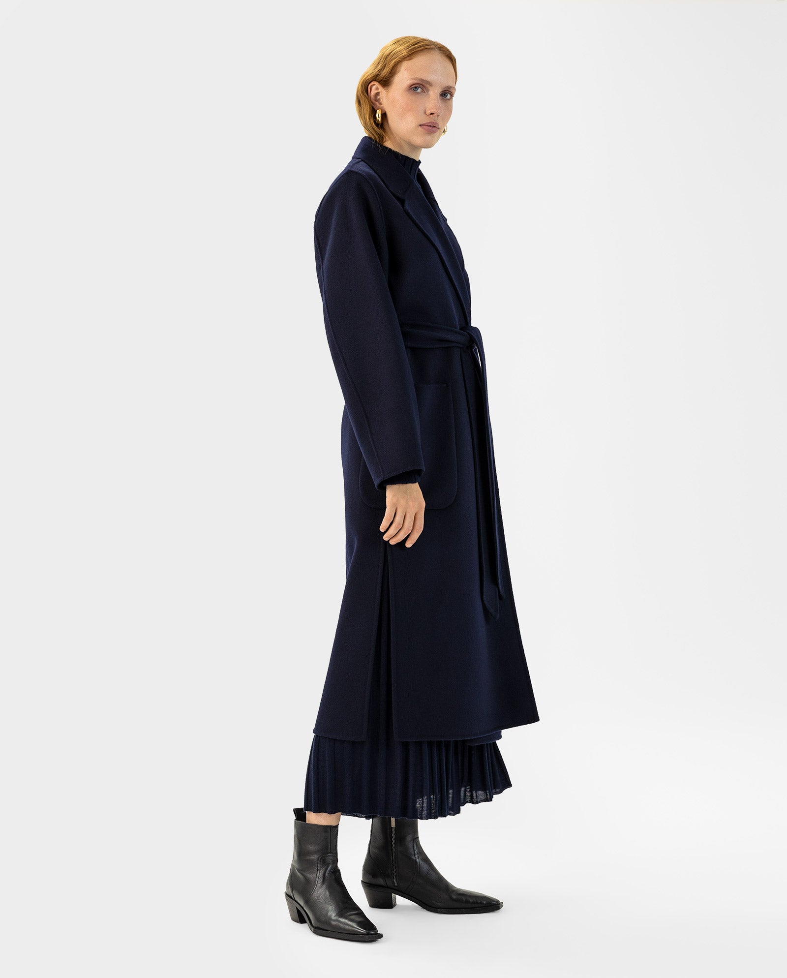 A woman wears the IVY OAK CELIA Doubleface Coat in navy wool, featuring a tie belt and midi length, paired with black boots and a skirt. Crafted to the Responsible Wool Standard, she stands against a white background.