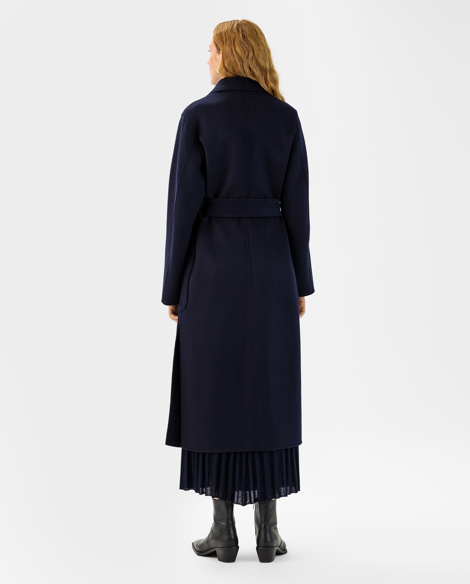 A woman stands with her back to the camera, wearing the IVY OAK CELIA Doubleface Coat in navy, paired with black boots and a pleated skirt.