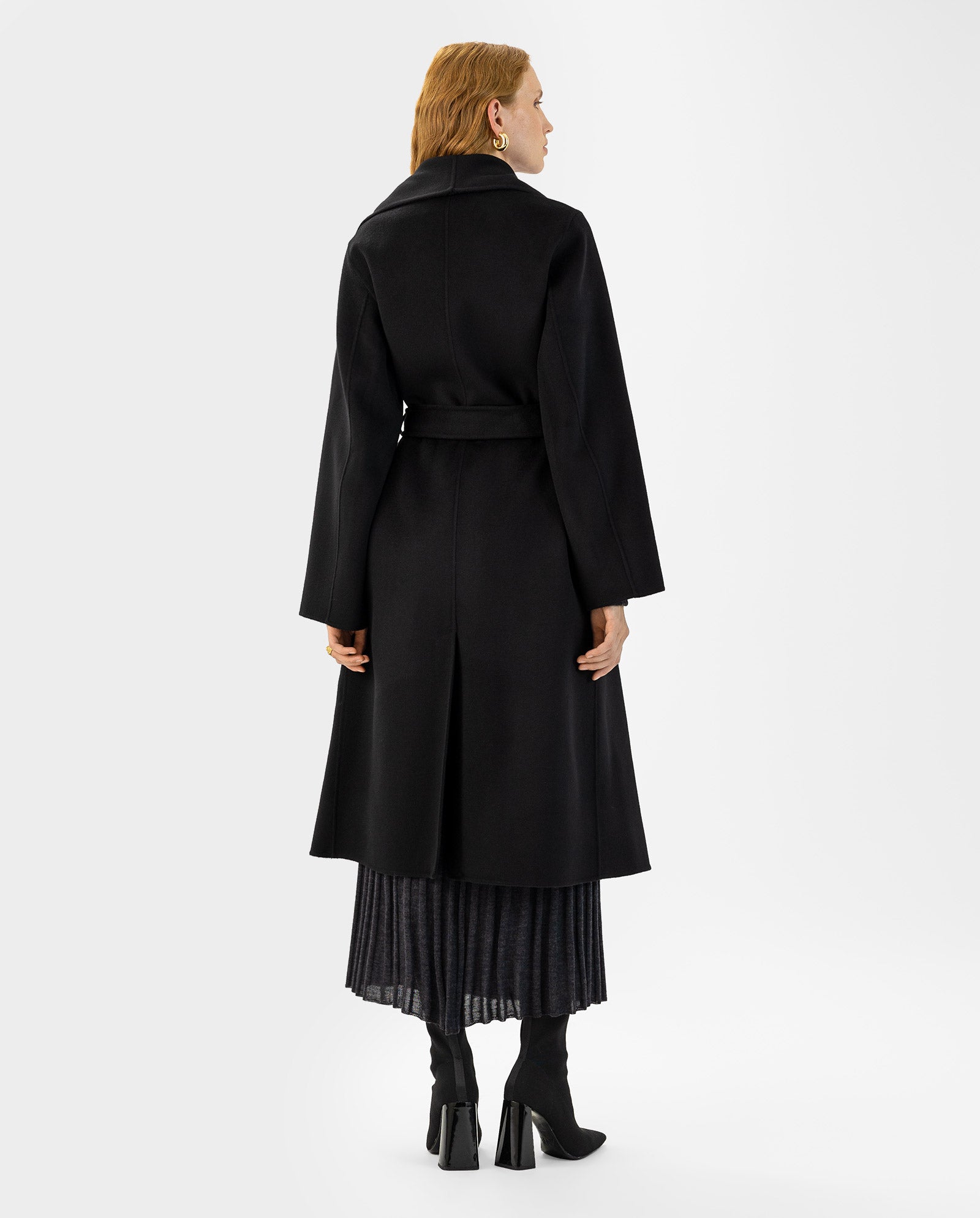 Minimalistic style: Woman in the IVY OAK CARRIE ROSE Wool Coat, black heeled boots, and pleated skirt stands with her back to the camera. The coat meets the Responsible Wool Standard for ethical fashion.