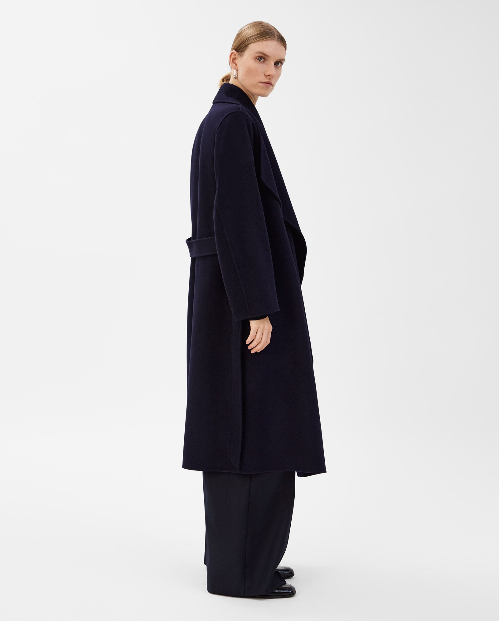 A woman stands sideways against a white background wearing the IVY OAK CARRIE ROSE Wool Coat, a long, dark navy minimalist piece with a big lapel collar and wide-leg pants. The coat is Responsible Wool Standard certified.