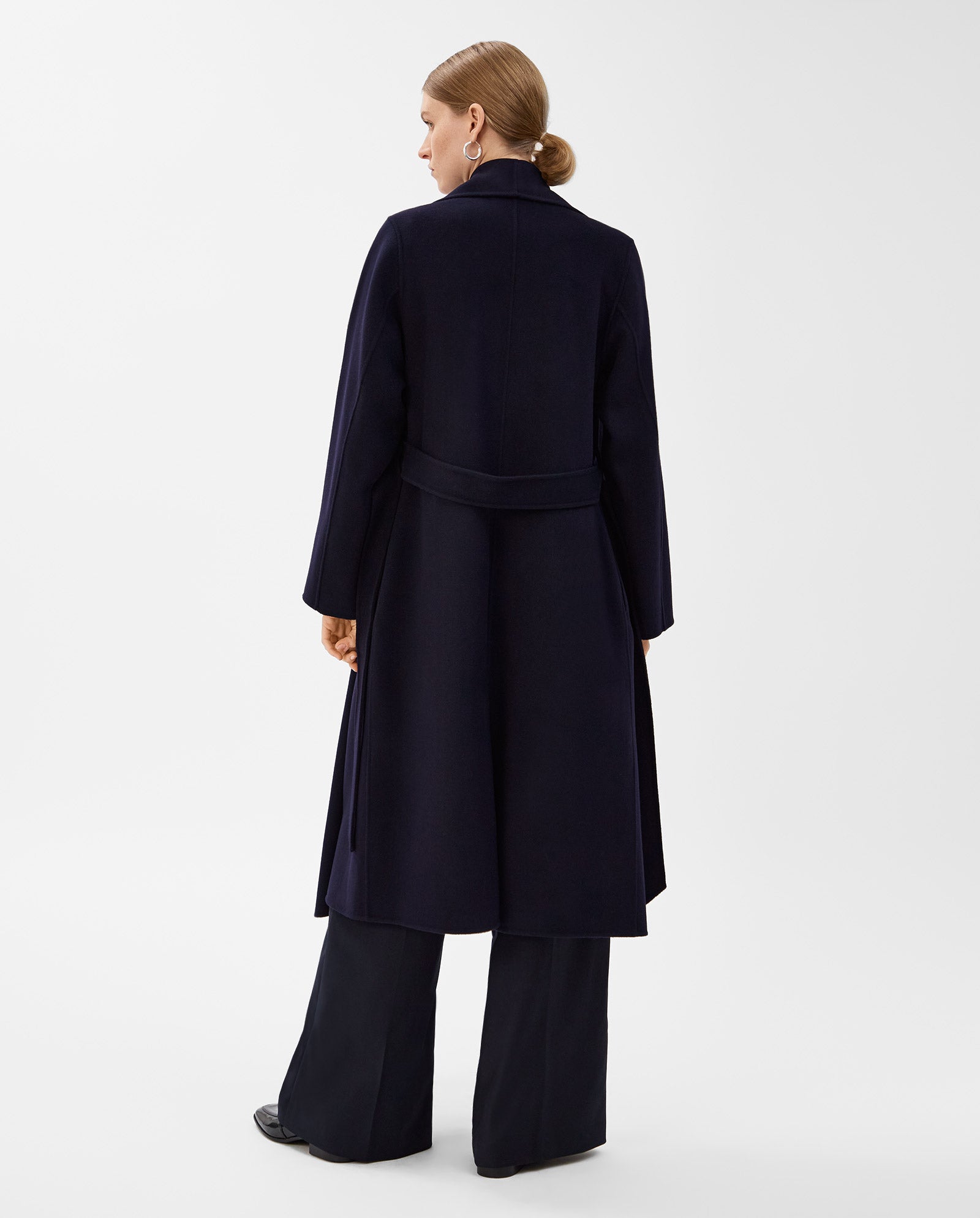 Woman facing away in the IVY OAK CARRIE ROSE Wool Coat—long, dark navy, minimalistic with a big lapel collar—paired with wide-leg pants. The coat is Responsible Wool Standard certified. White background.