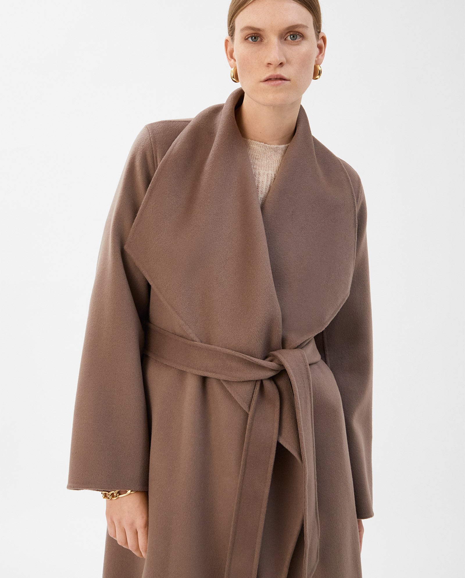 A woman wears the IVY OAK CARRIE ROSE Wool Coat, a minimalist taupe design with wide lapels and a belt, crafted in line with the Responsible Wool Standard. She stands against a plain white background.