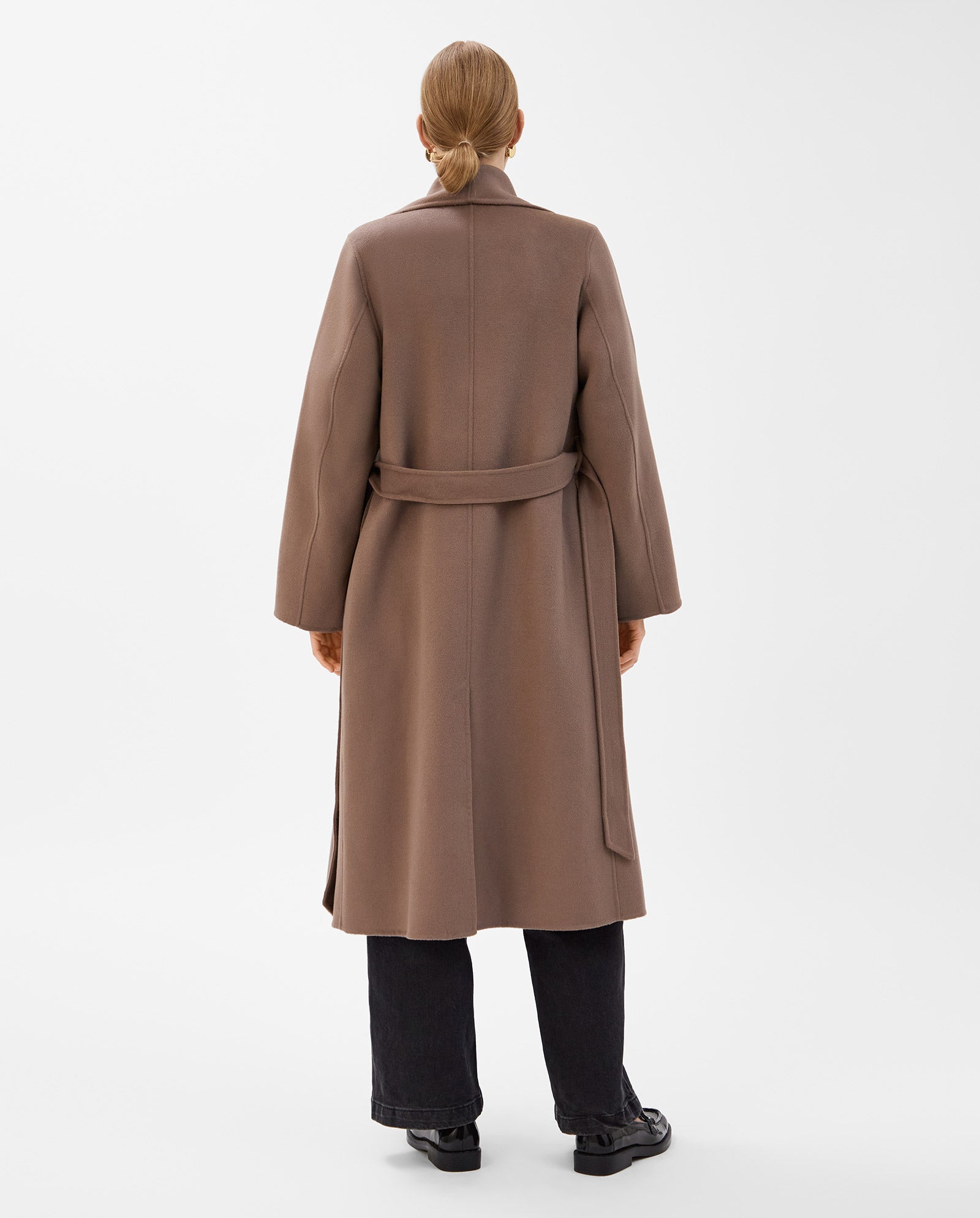 A minimalist scene features a person with tied-back hair in the IVY OAK CARRIE ROSE Wool Coat, crafted from Responsible Wool Standard-certified brown wool, and black pants, seen from behind.