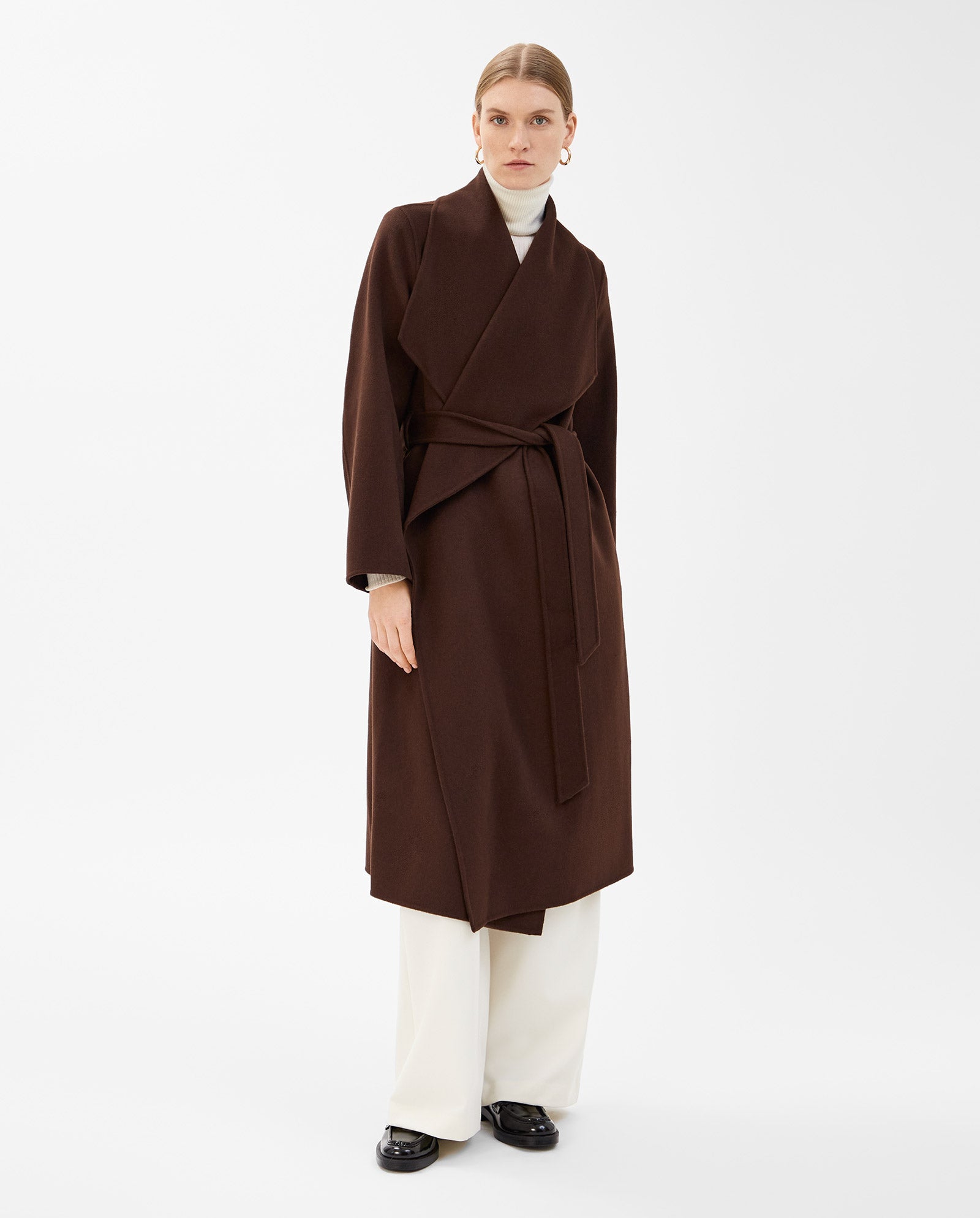 A woman wears the IVY OAK CARRIE ROSE Wool Coat, a minimalistic long brown piece certified by the Responsible Wool Standard, styled over a white turtleneck, wide-leg white pants, and black shoes.
