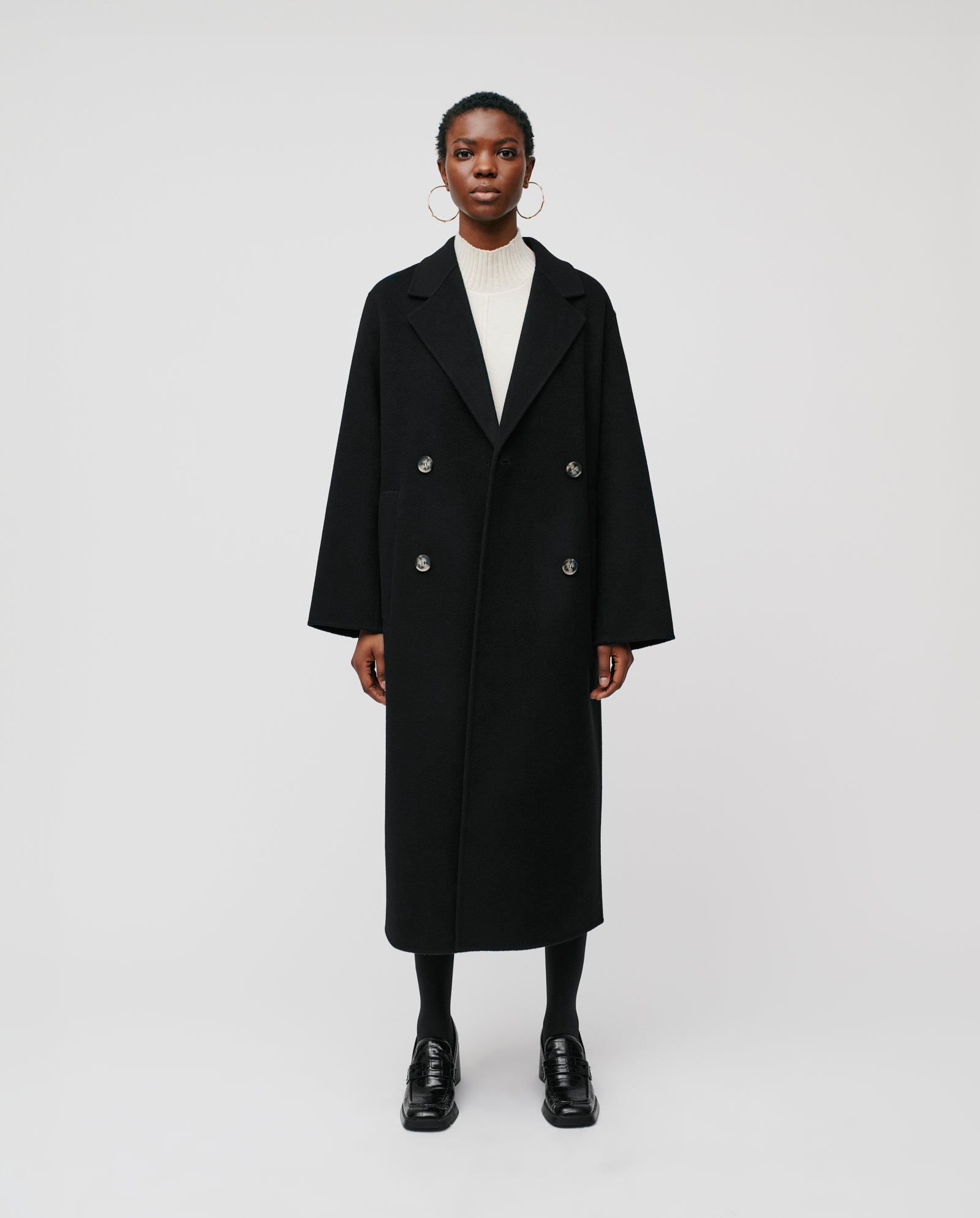 A person stands facing forward wearing the IVY OAK CLARA Wool Coat—a long black double-breasted, oversized midi coat—paired with a white sweater, black tights, and black shoes.