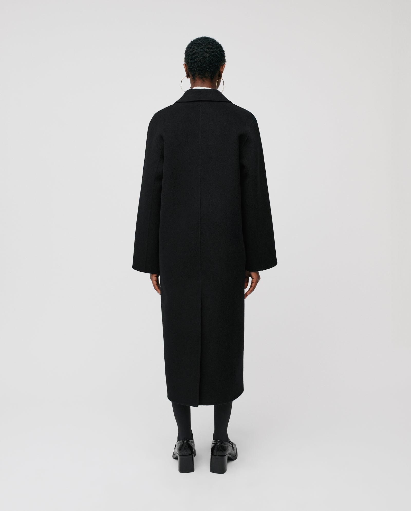 Model wearing the IVY OAK CLARA Wool Coat, a long black midi coat with an oversized fit, paired with boots and standing against a plain white background, facing away.