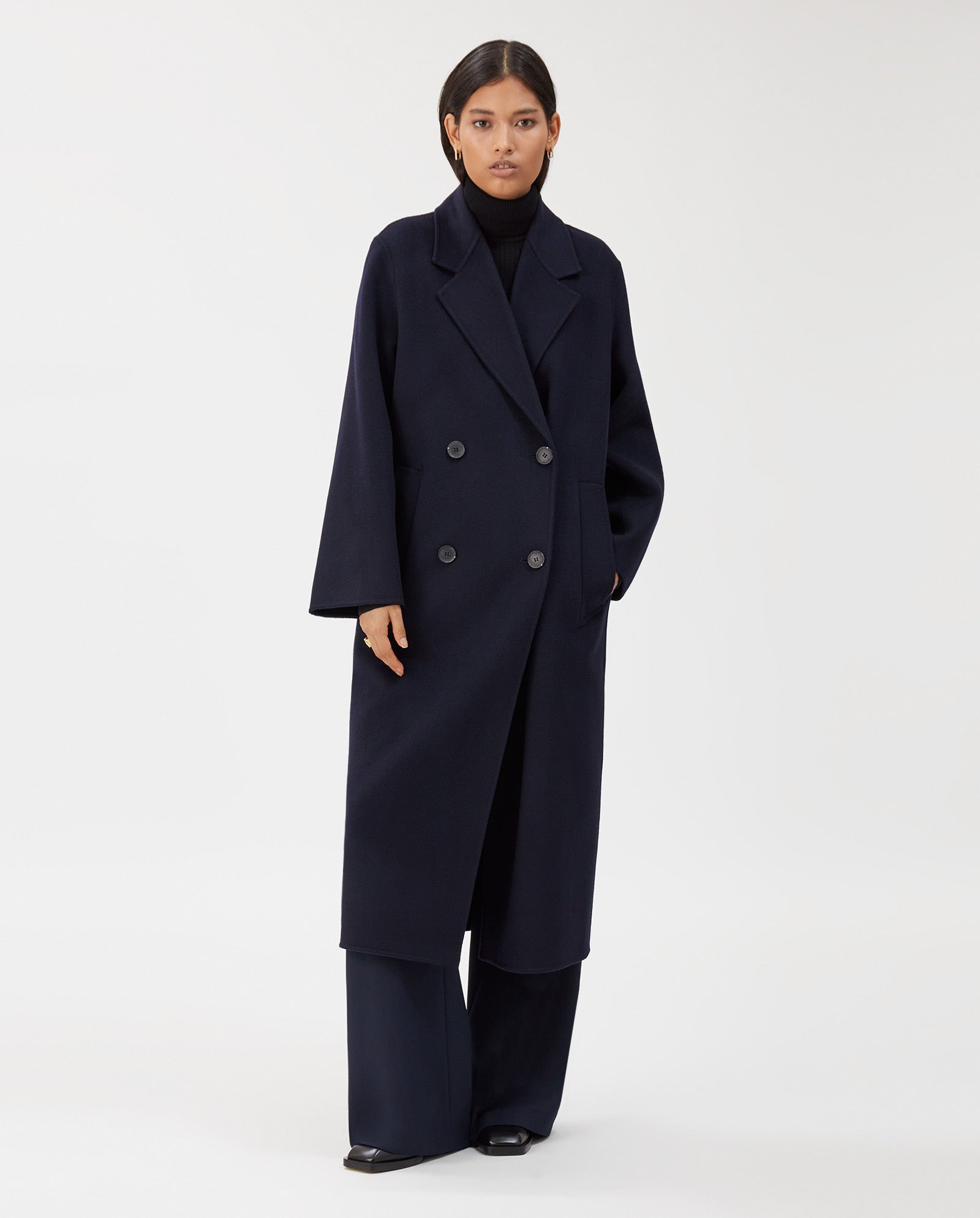 A woman wearing the IVY OAK CLARA Wool Coat—a long, dark navy midi coat with an oversized fit—and wide-leg pants stands against a white background.