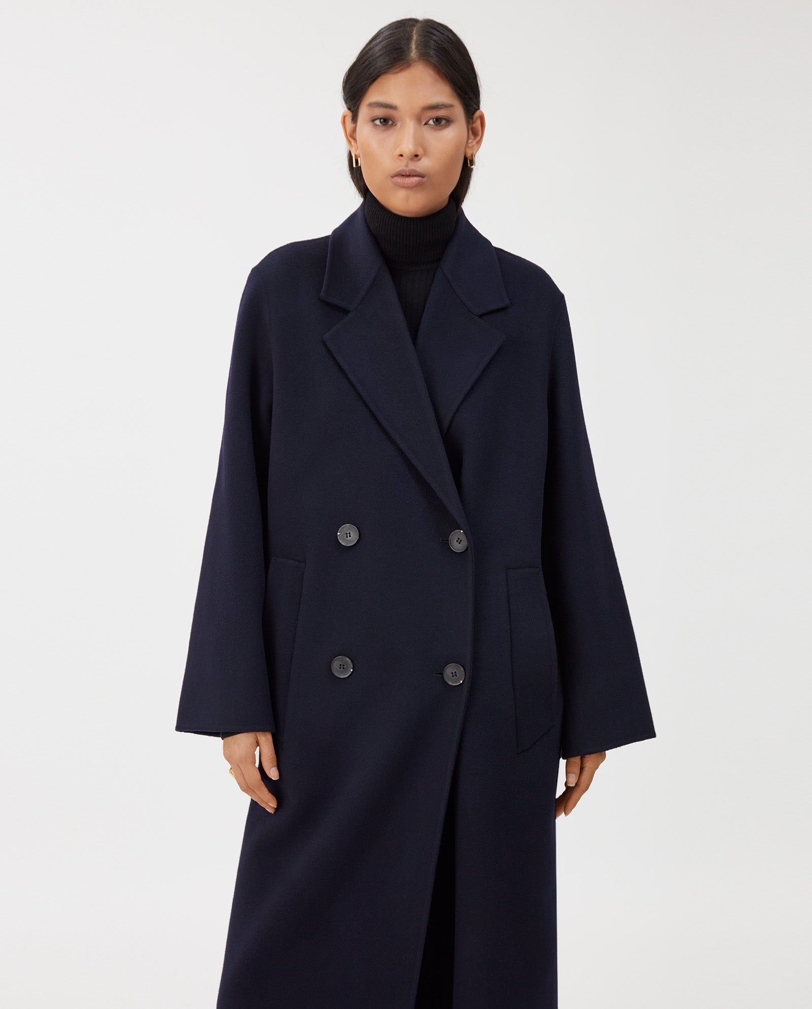 A woman wears the IVY OAK CLARA Wool Coat—an oversized, dark navy, double-breasted midi coat made to the Responsible Wool Standard—over a black turtleneck, standing before a white background.