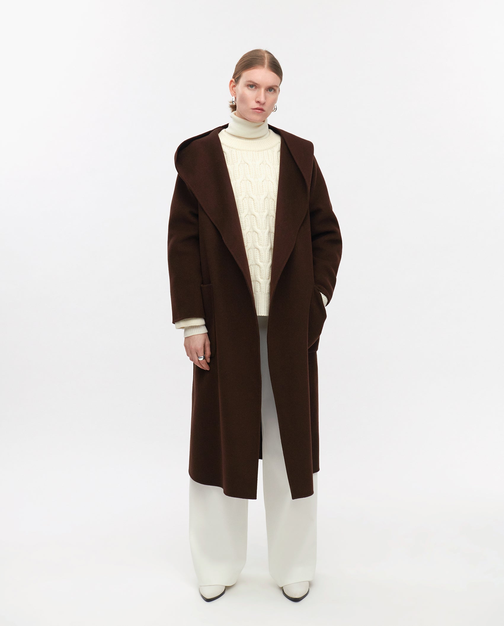 A person wears the IVY OAK CELIA EDIE dark brown wool coat, certified by the Responsible Wool Standard, paired with a white turtleneck and white pants against a plain background.