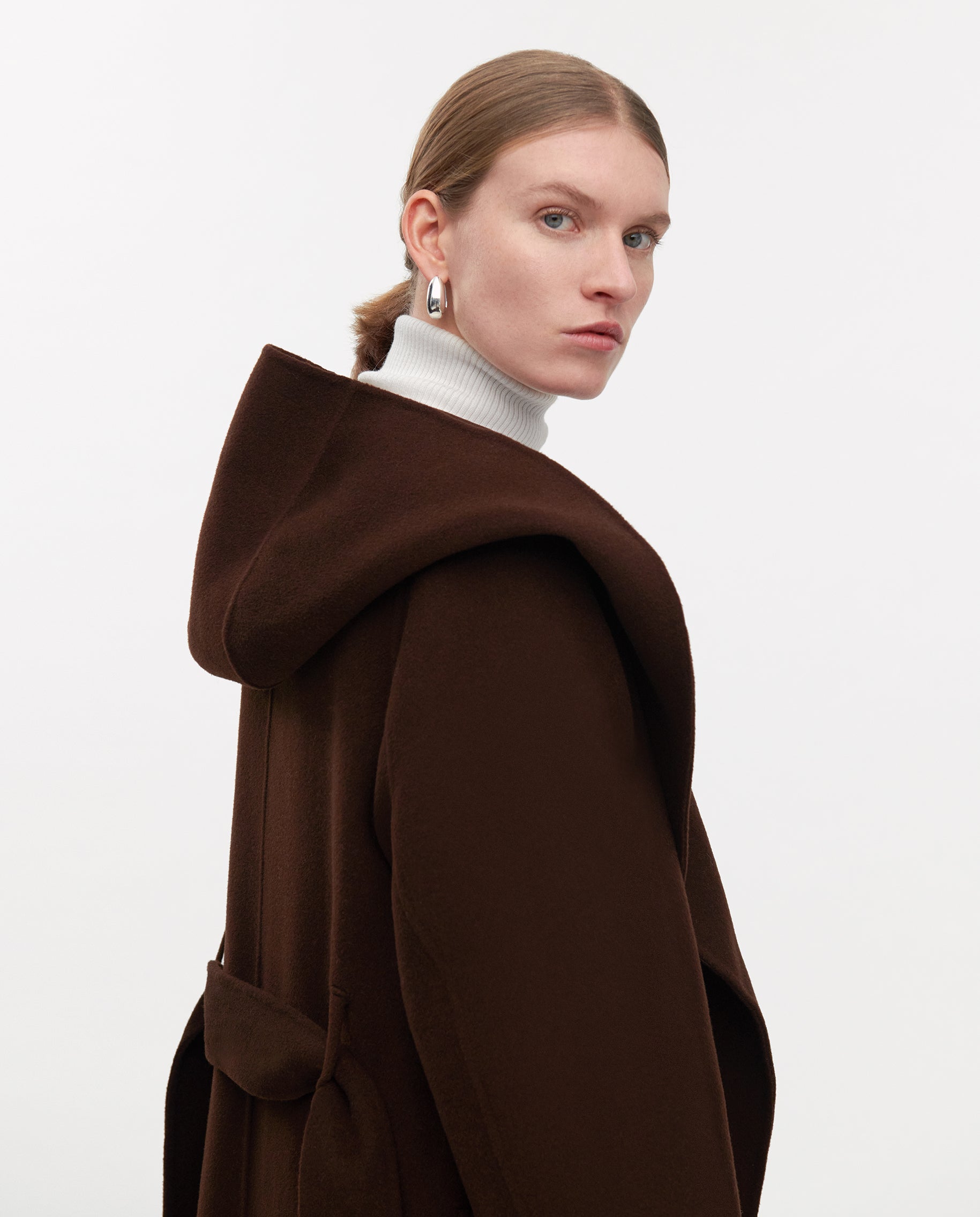 Woman wearing the IVY OAK CELIA EDIE Coat, made from Responsible Wool Standard-certified brown wool, layered over a white turtleneck and looking over her shoulder against a plain background.