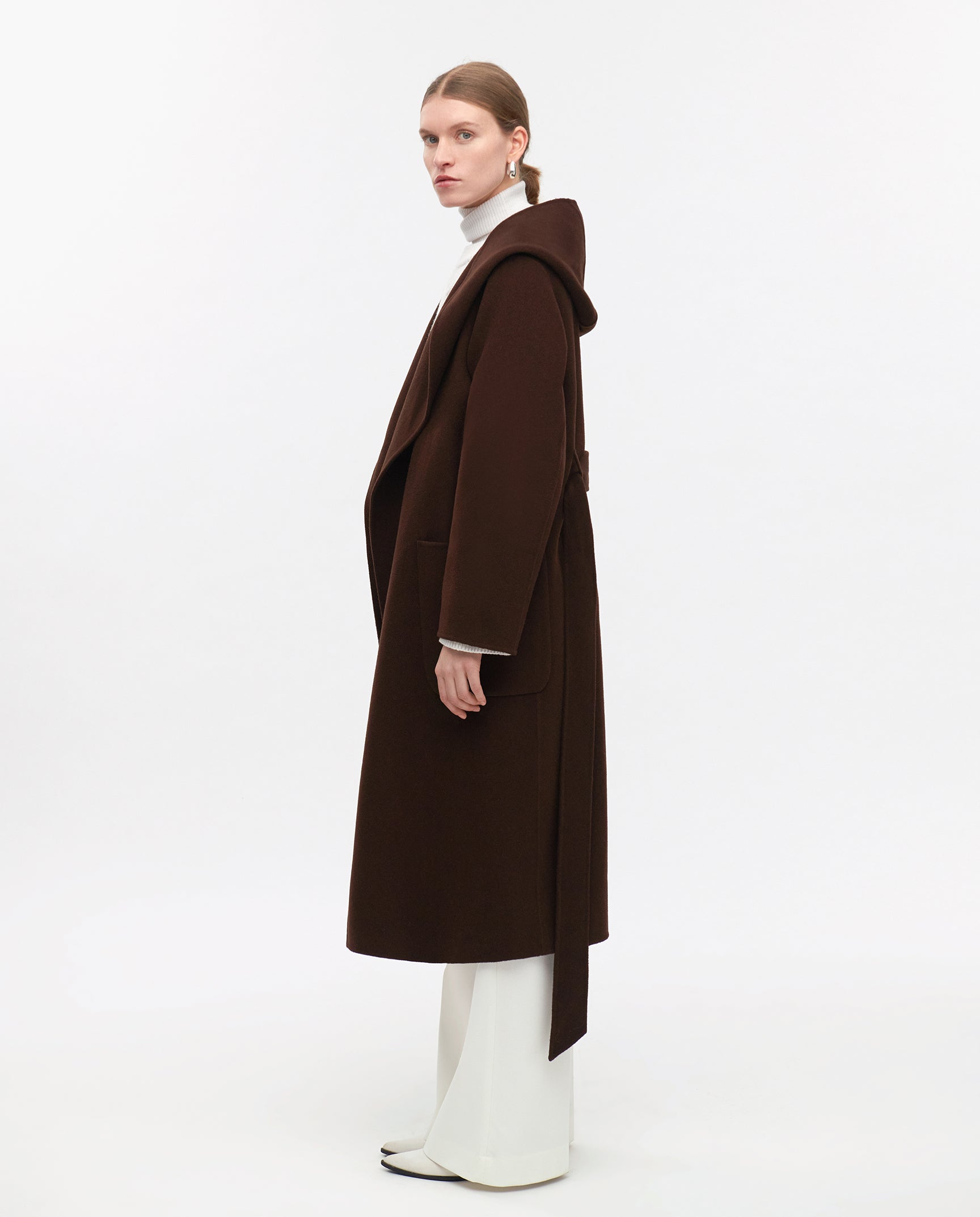 A woman models the CELIA EDIE Coat by IVY OAK, a long dark brown woolcoat certified by the Responsible Wool Standard, layered over a white turtleneck and pants against a plain background.