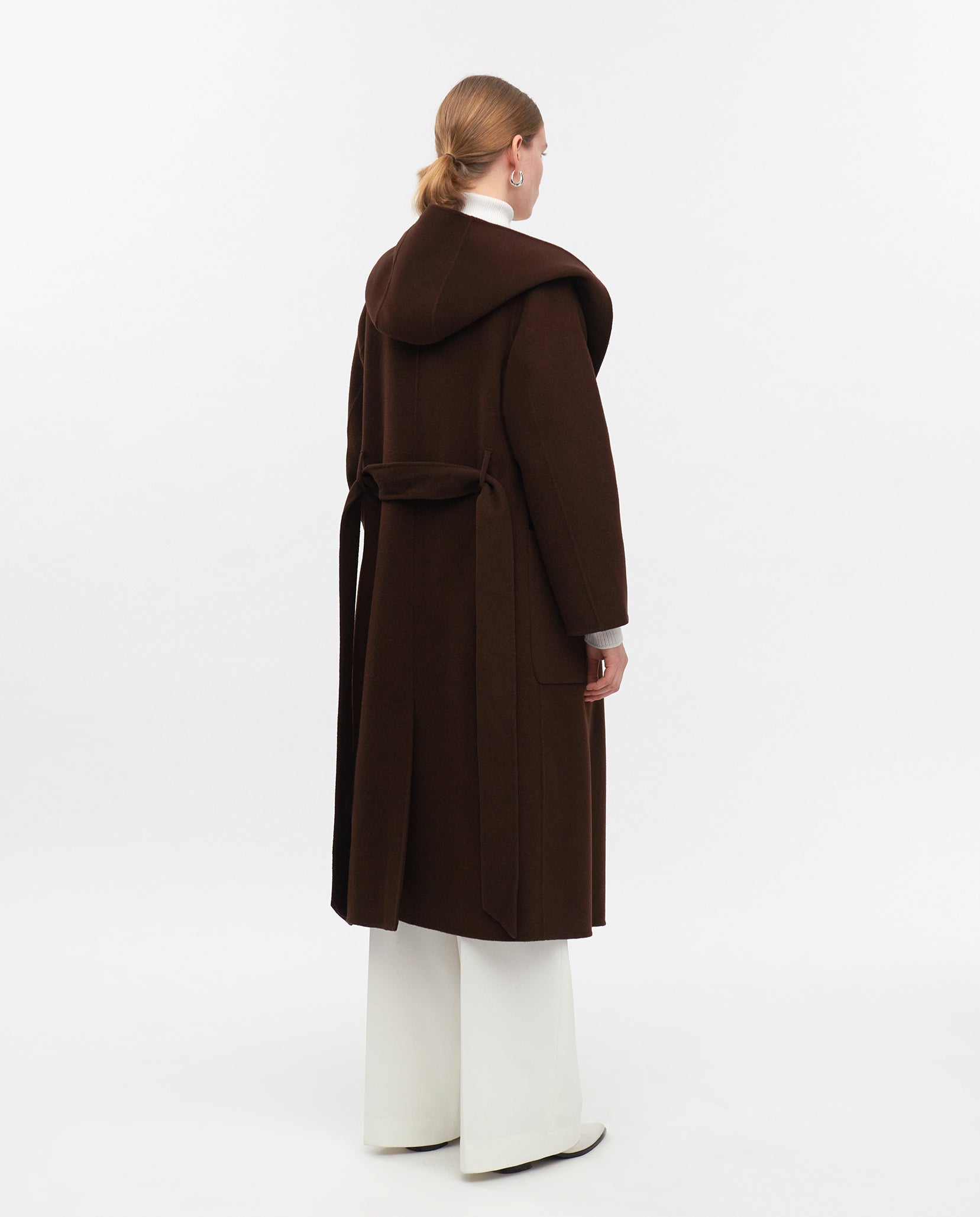 A woman wears the IVY OAK CELIA EDIE long dark brown wool coat with a hood, RWS certified, styled with white pants and black shoes, standing against a white background.