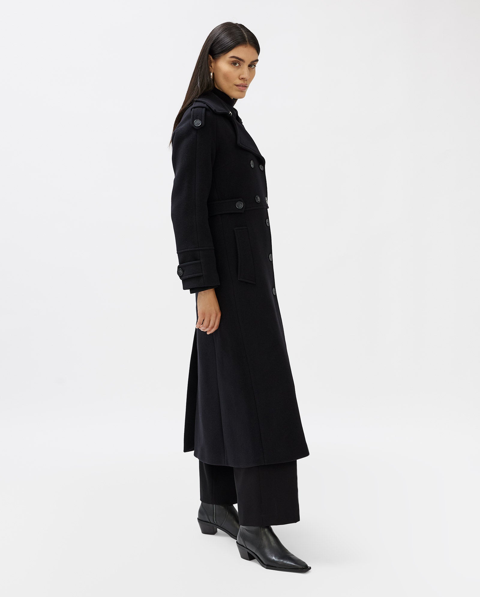 A woman wearing the IVY OAK CANDELA long black wool midi coat with shoulder pads, paired with black pants and boots, stands against a white background.