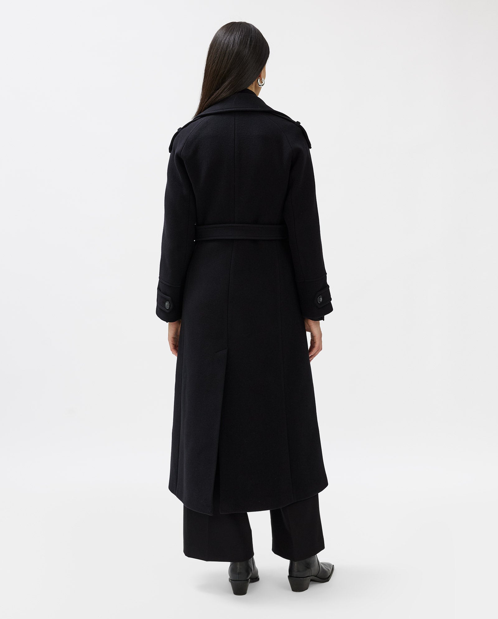 A woman stands with her back to the camera in IVY OAKs CANDELA Coat, a wool midi style with shoulder pads, paired with black pants against a white background.
