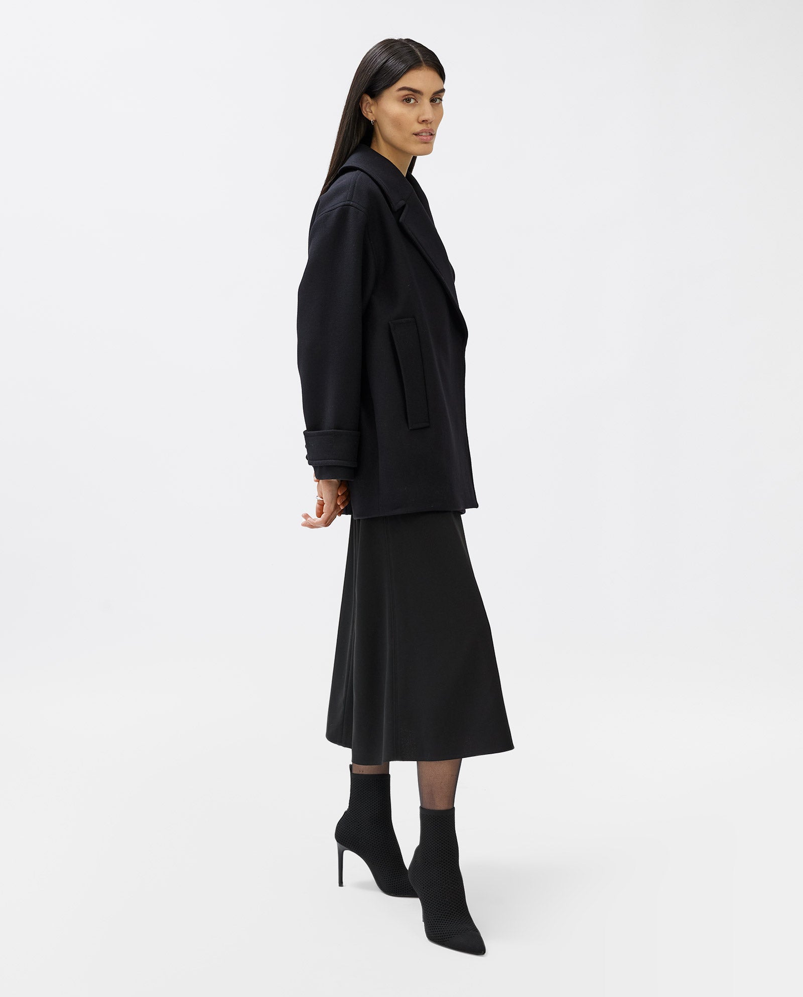 A woman wears the IVY OAK CARLY Coat, a loose fit black design with hidden button placket, styled with a midi skirt and heeled boots against a plain white background.