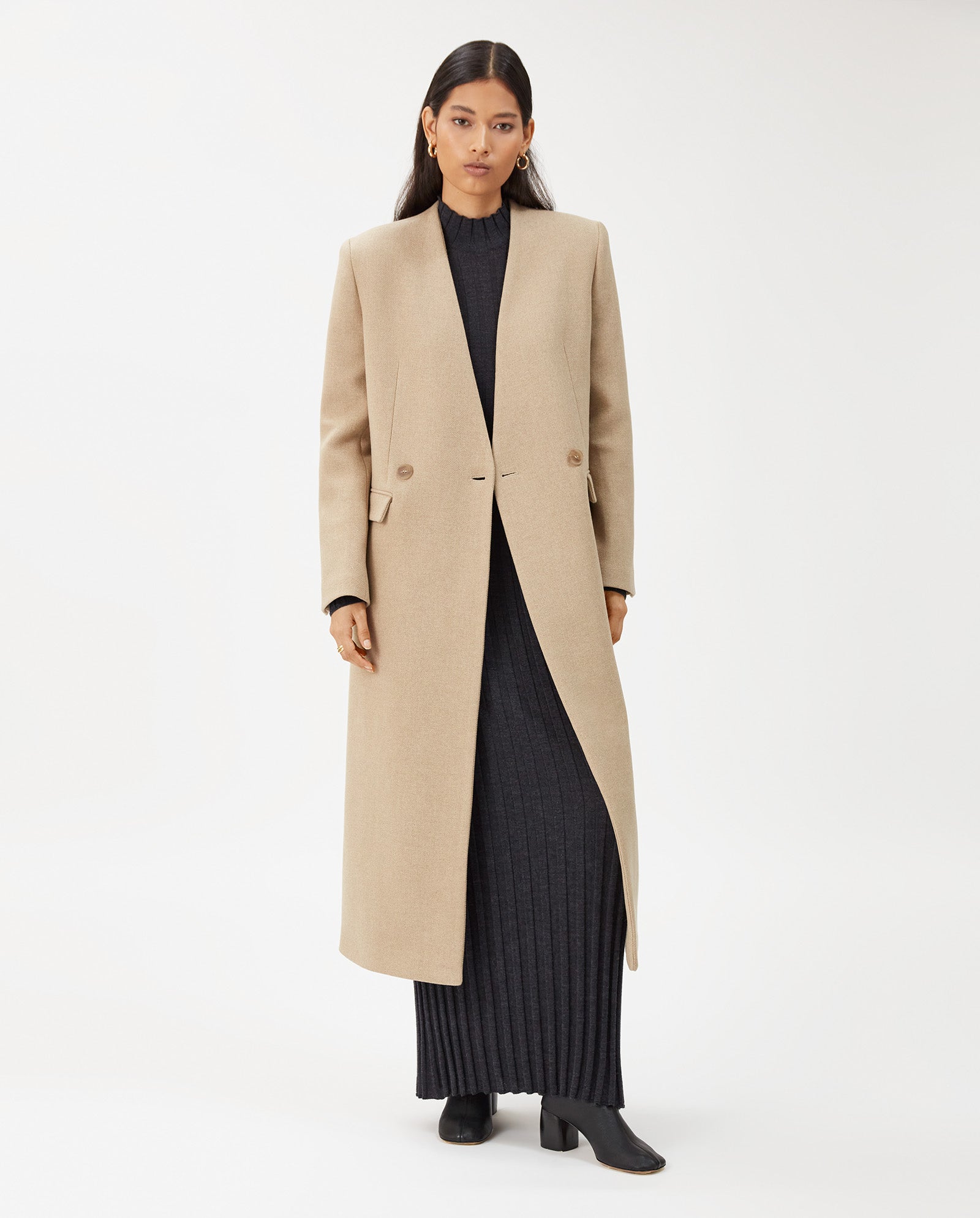 A woman wears the IVY OAK CAPRI coat, a maxi-length beige wool style with a stand-up collar, layered over a black ribbed dress and black boots, standing against a plain background.