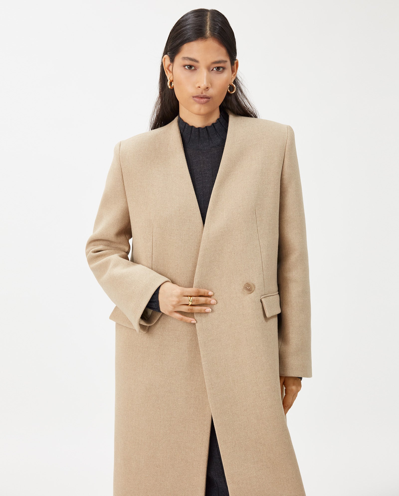 A woman wearing the IVY OAK CAPRI Coat, a beige wool design with a stand-up collar over a dark top, stands against a plain white background.