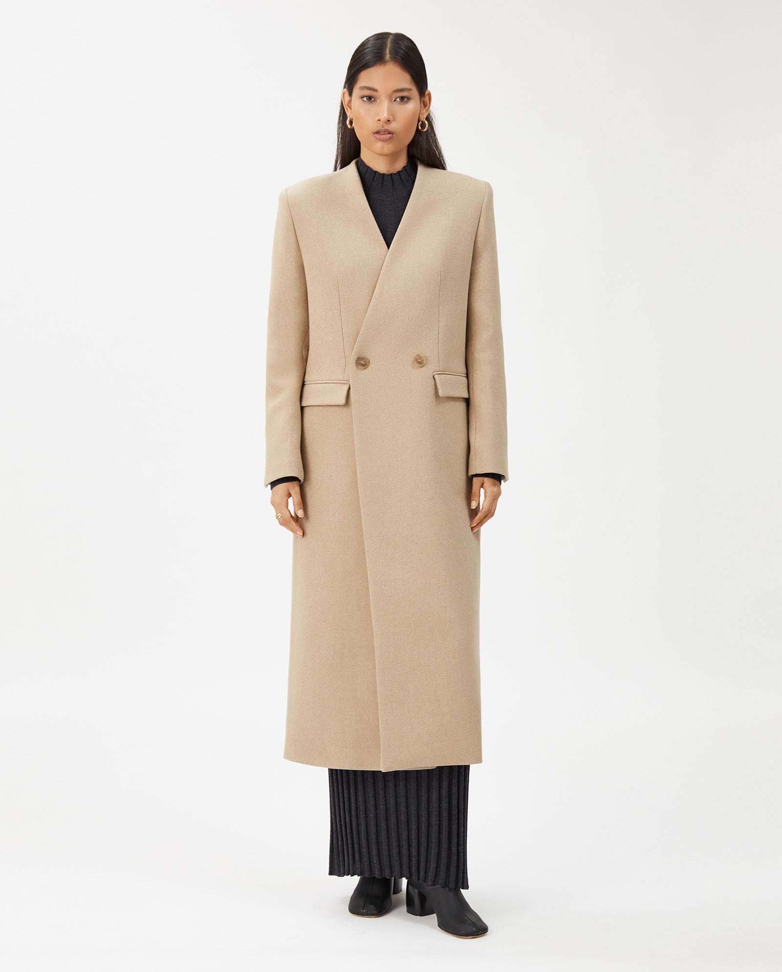 A woman stands facing forward wearing the IVY OAK CAPRI Coat, a beige wool maxi-length style with a stand-up collar, over a black ribbed dress and black shoes.