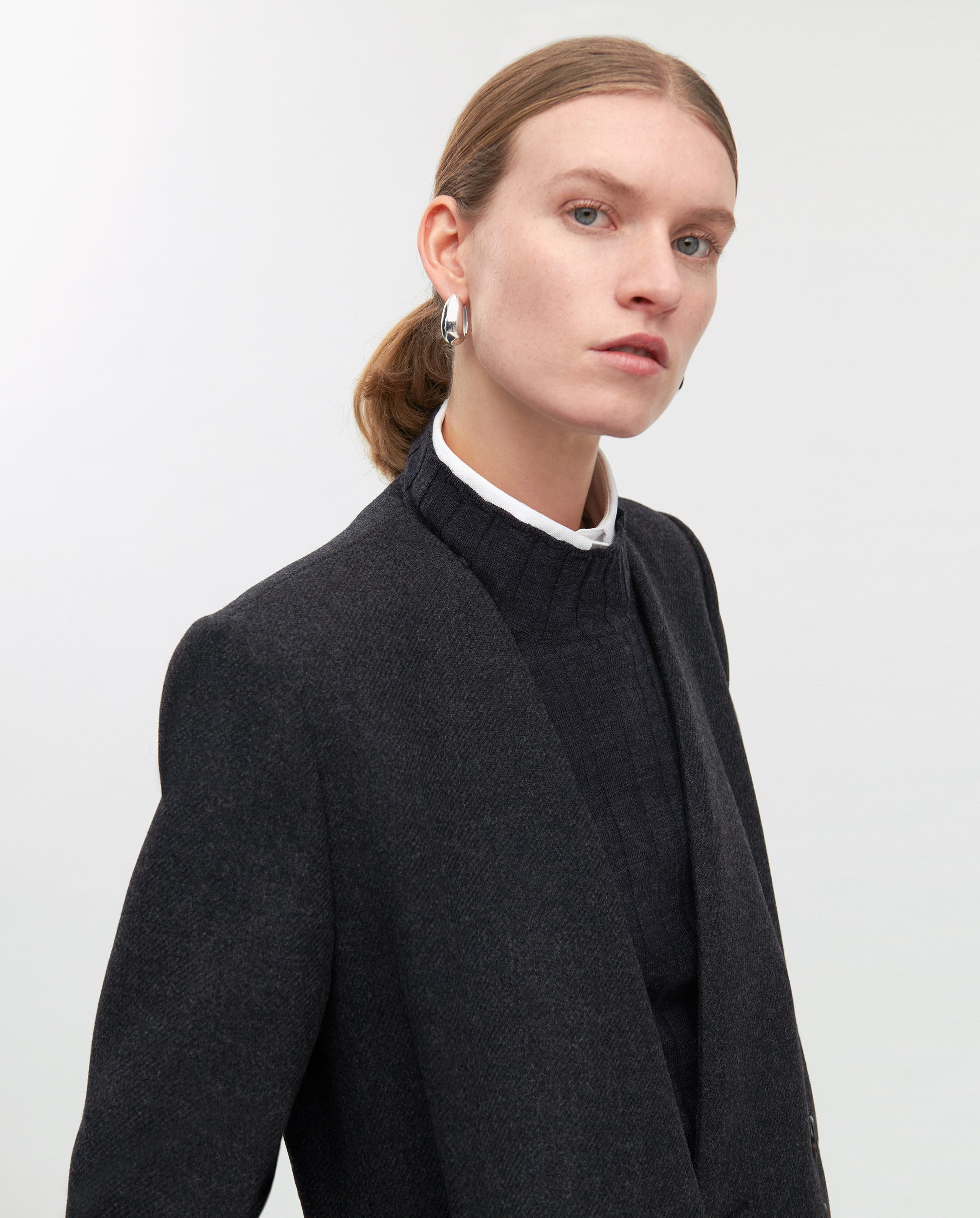 Woman with slicked-back hair wears a dark blazer, black top, silver hoop earrings, and the elegant IVY OAK CAPRI Coat, maxi length wool outerwear, posed against a plain background.