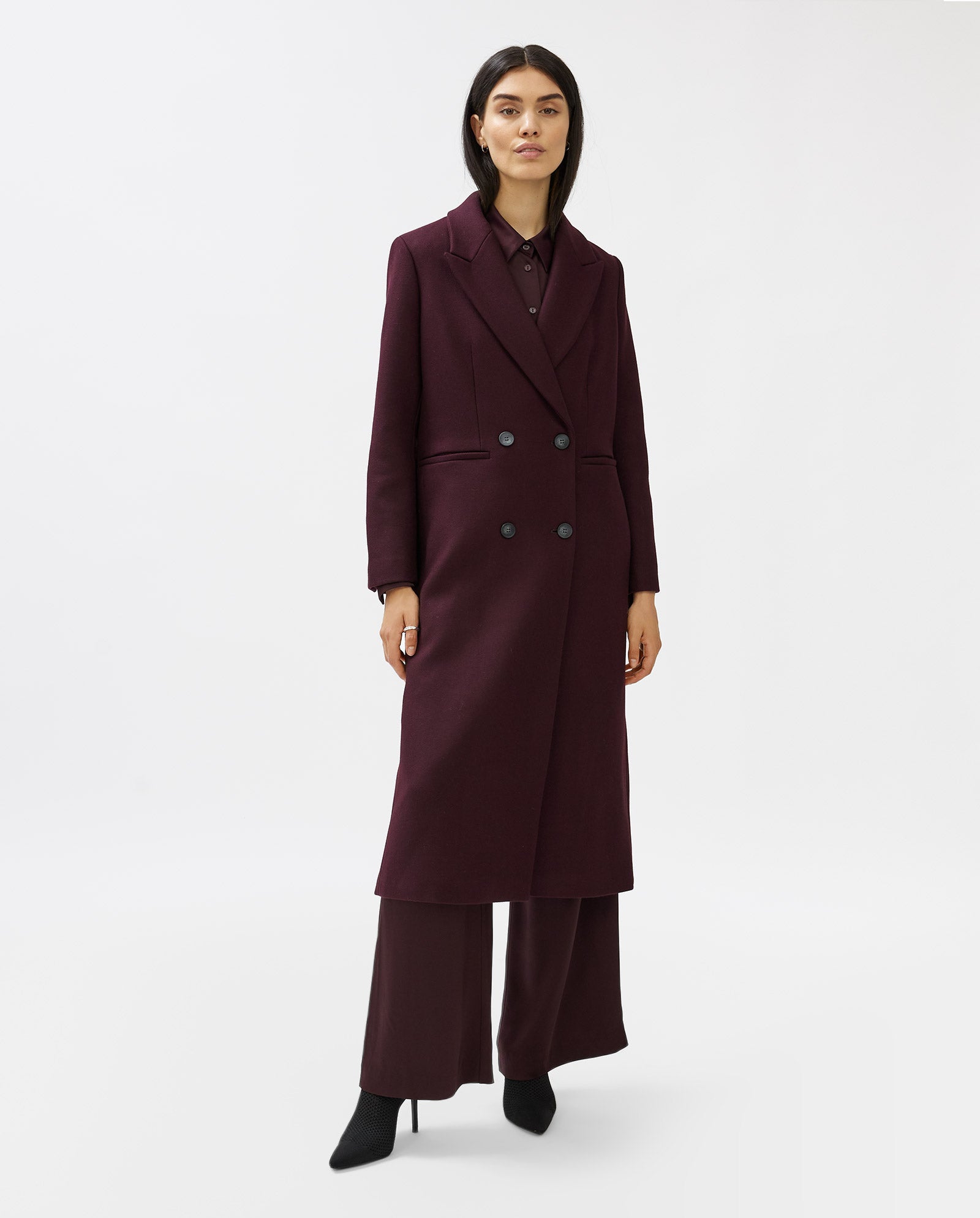 A woman stands against a white background wearing the IVY OAK CAYENNE coat—a long, dark burgundy knee-length wool piece—paired with matching pants and black shoes.