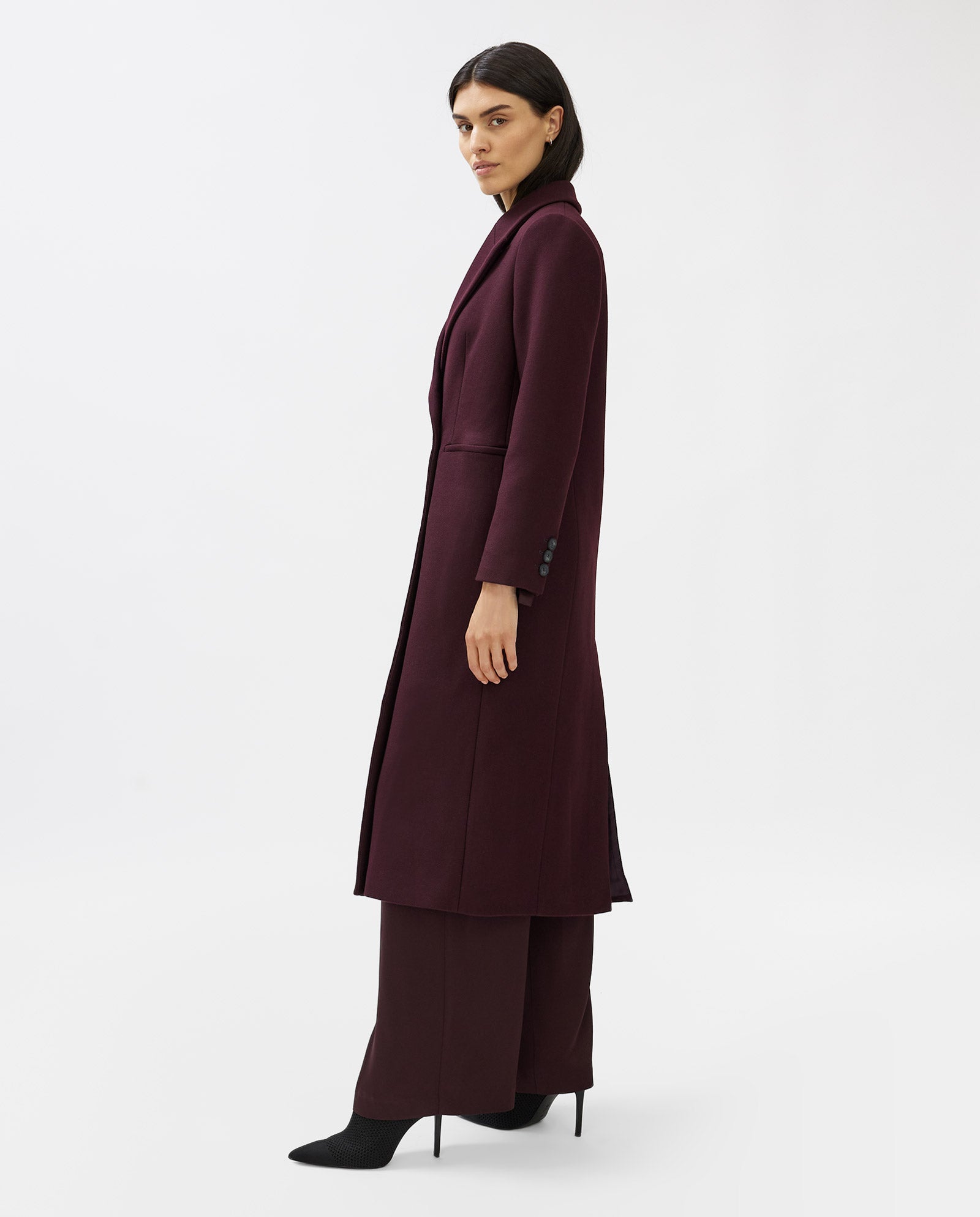 A woman wears the IVY OAK CAYENNE coat, a loose-fit, knee-length burgundy piece, paired with black heels. She stands against a plain white background, gazing to the side.