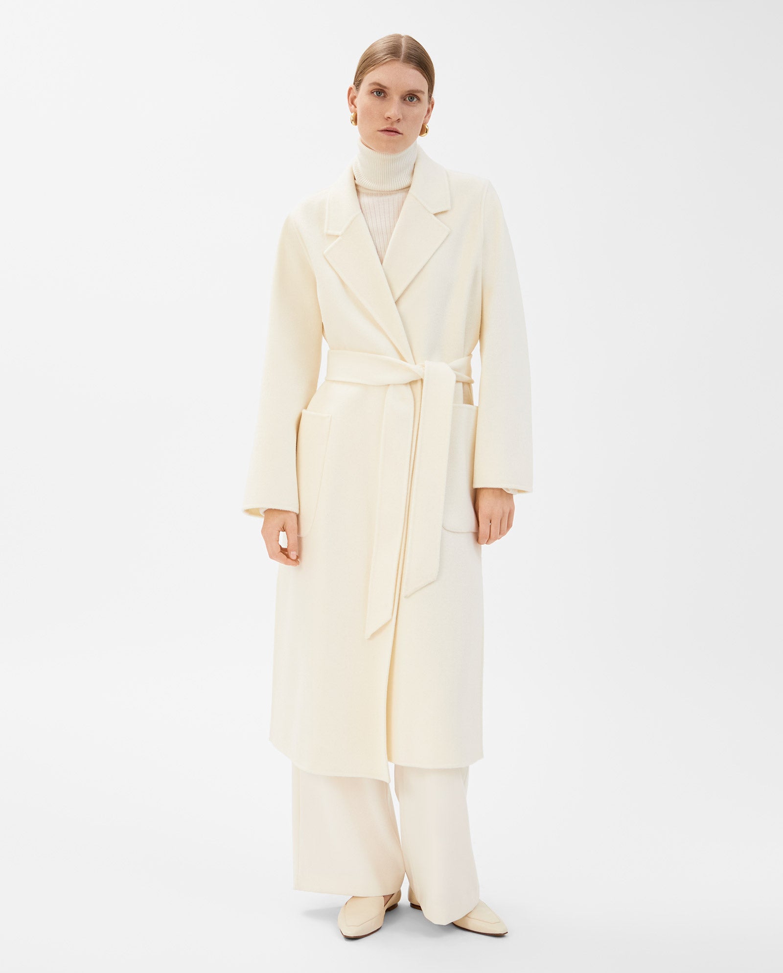 A woman wears the IVY OAK CELIA Doubleface Wool Coat, a cream-colored, belted wool coat, styled with a turtleneck, loose wide-leg pants, and cream shoes against a white background.