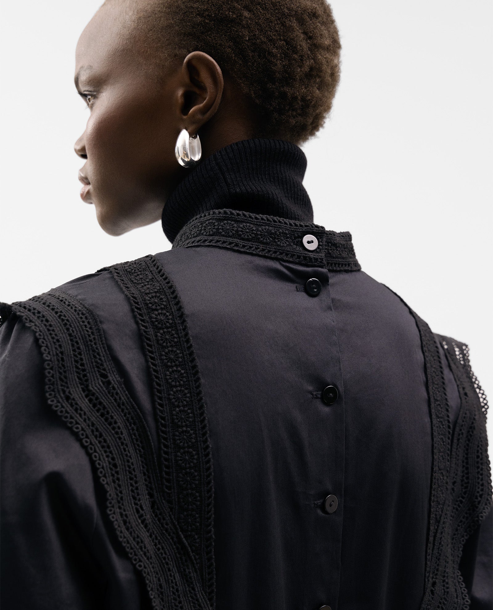 A woman wears the IVY OAK ELINOR Blouse, a black cotton top featuring a buttoned back, lace tapes, and cropped fit, styled with silver hoop earrings.