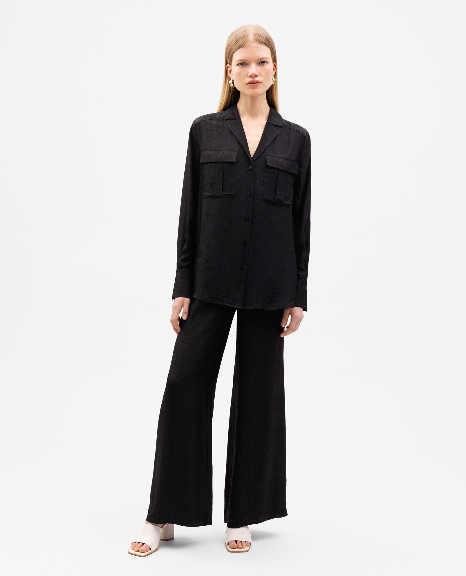 A woman wears the IVY OAK EDITH Blouse, an oversized black top with a button placket and front pockets, paired with matching wide-leg pants, standing against a white background.