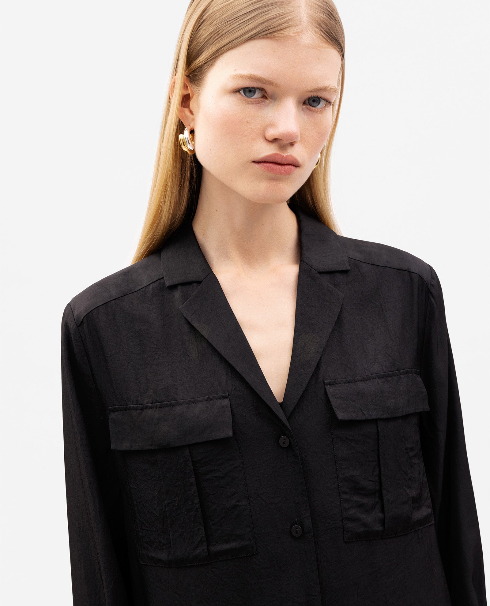 Woman with straight blonde hair wearing the IVY OAK EDITH Blouse—a black, shiny top featuring front pockets and a button placket—looking at the camera.