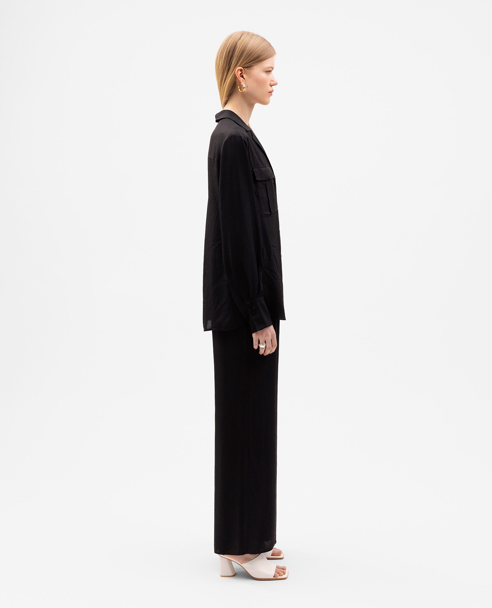 A woman in the IVY OAK EDITH Blouse—an oversized black top with a button placket—paired with matching pants and white heels, is shown in side view against a white background.