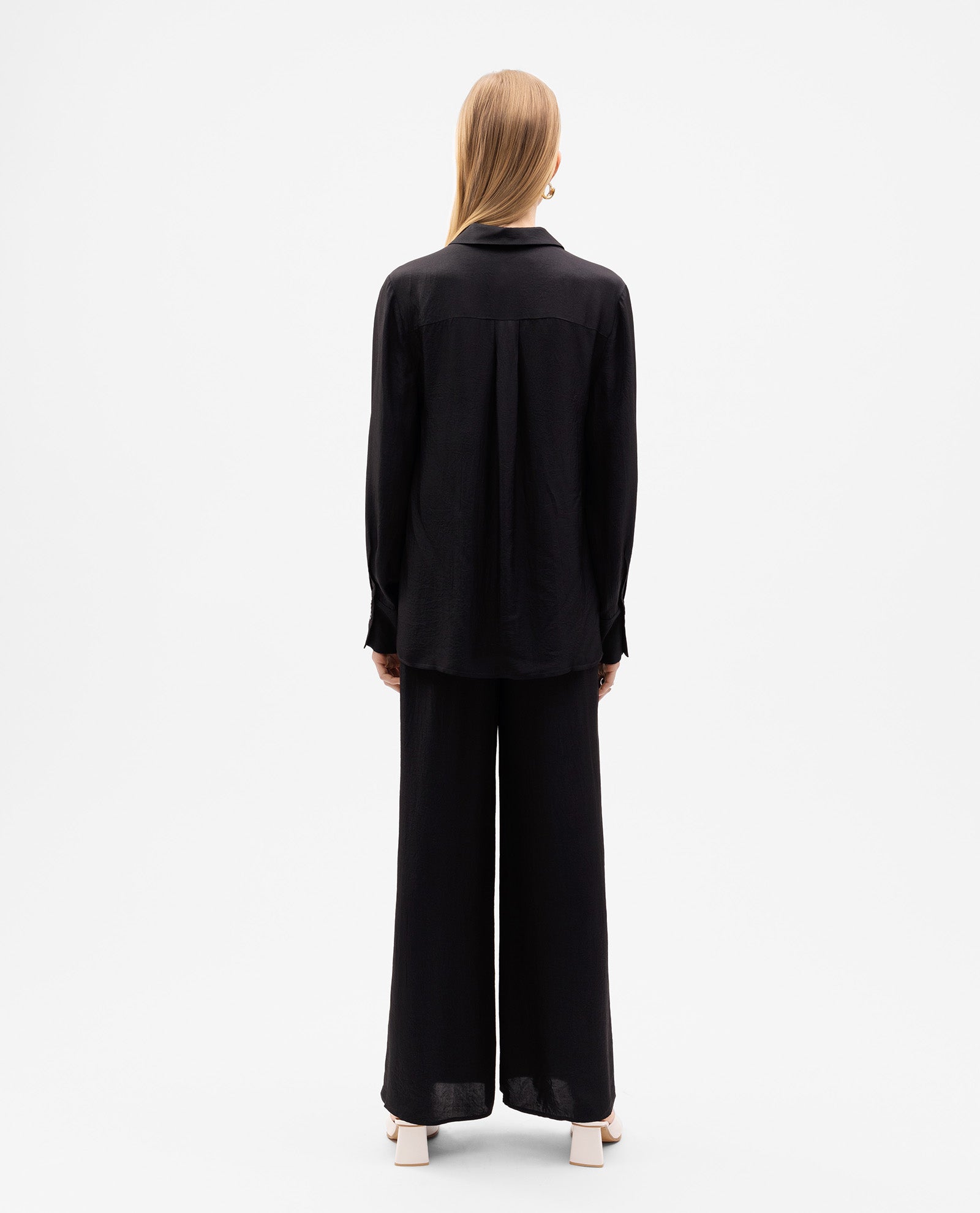 Person with straight blonde hair, facing away, wears the oversized black EDITH Blouse by IVY OAK with a button placket and wide-leg pants.