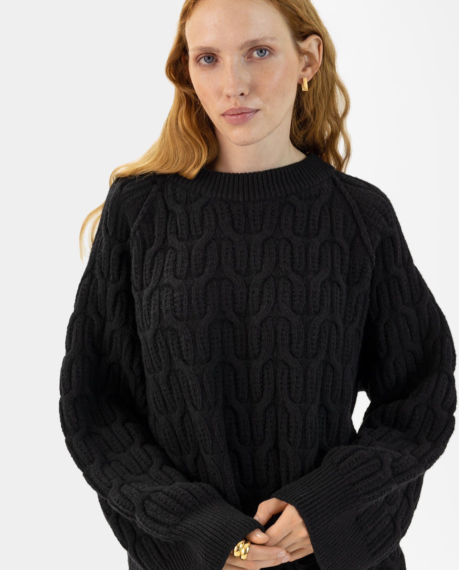 A woman with long red hair wears the IVY OAK KLARINA Jumper, a wide, loose-fit textured black soft knit, paired with gold earrings and a gold ring, and stands against a white background.