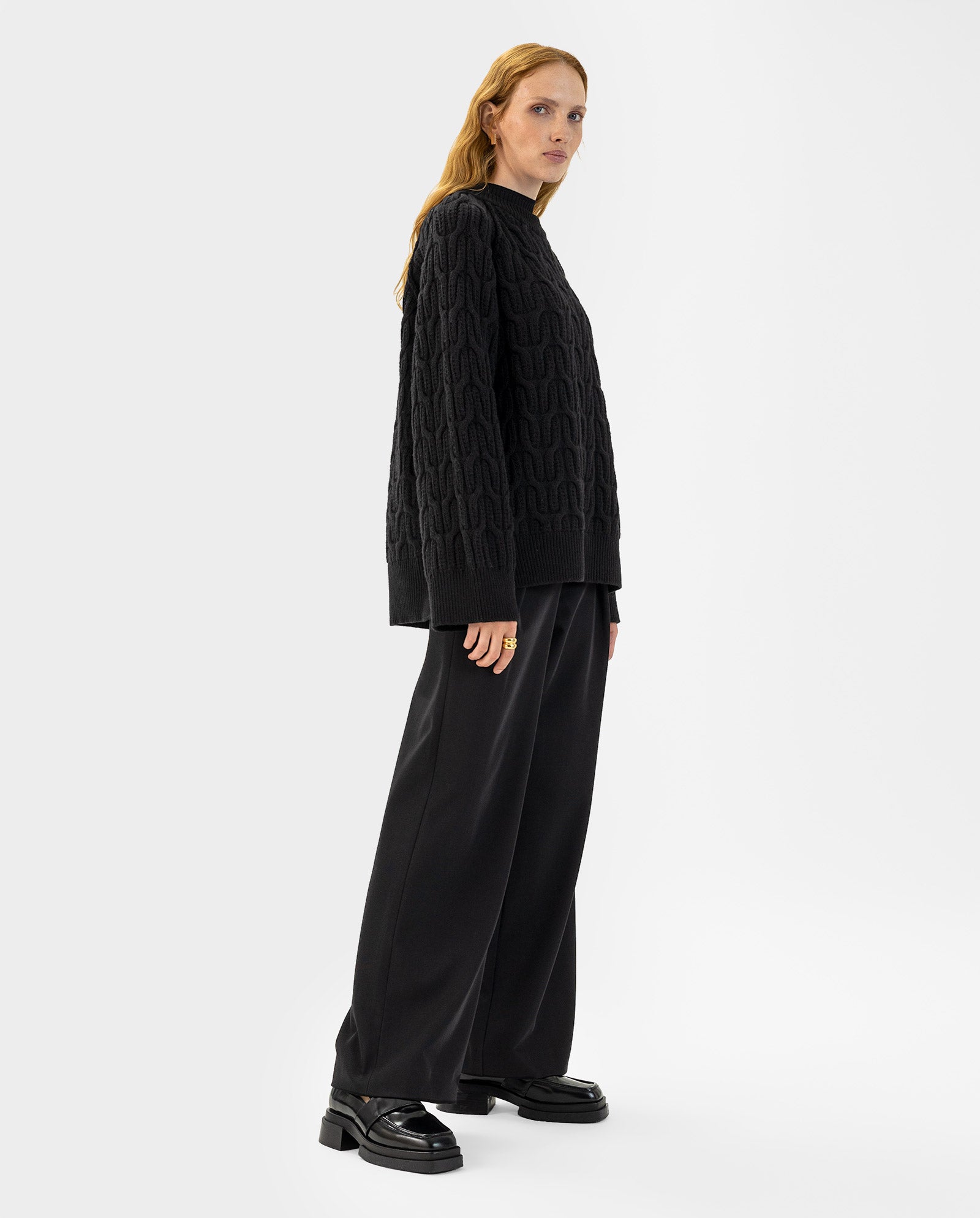 A woman stands sideways against a plain white background, wearing the IVY OAK KLARINA Jumper in black—a soft knit with a wide, loose fit—paired with wide-leg pants.