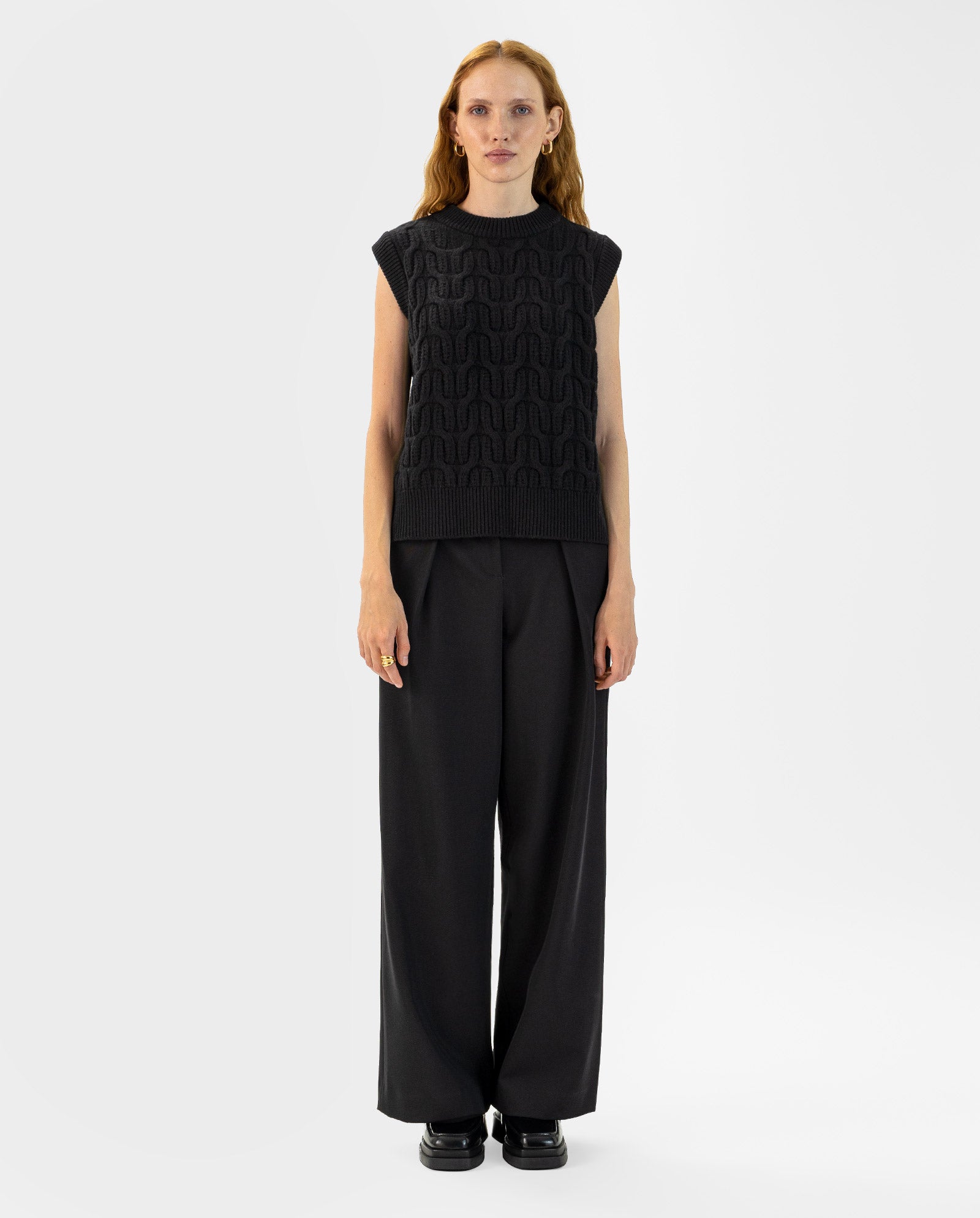 Woman with long red hair wears the IVY OAK KLARISSA Pullunder, a black boxy fit sleeveless top, styled with black wide-leg pants against a white background.