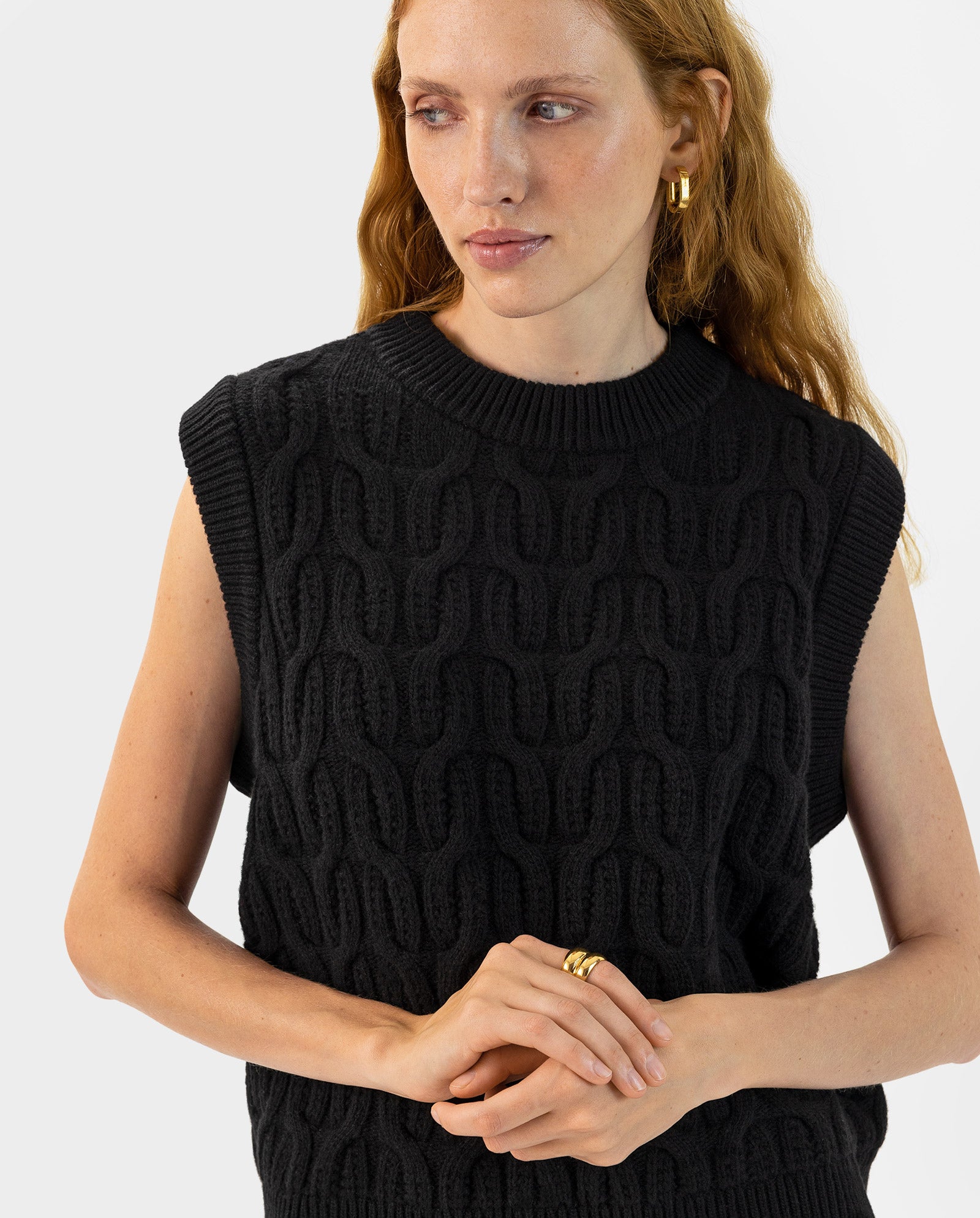 A woman with long red hair wears the IVY OAK KLARISSA Pullunder, a sleeveless black textured knit in a boxy fit, styled with gold hoop earrings and rings.