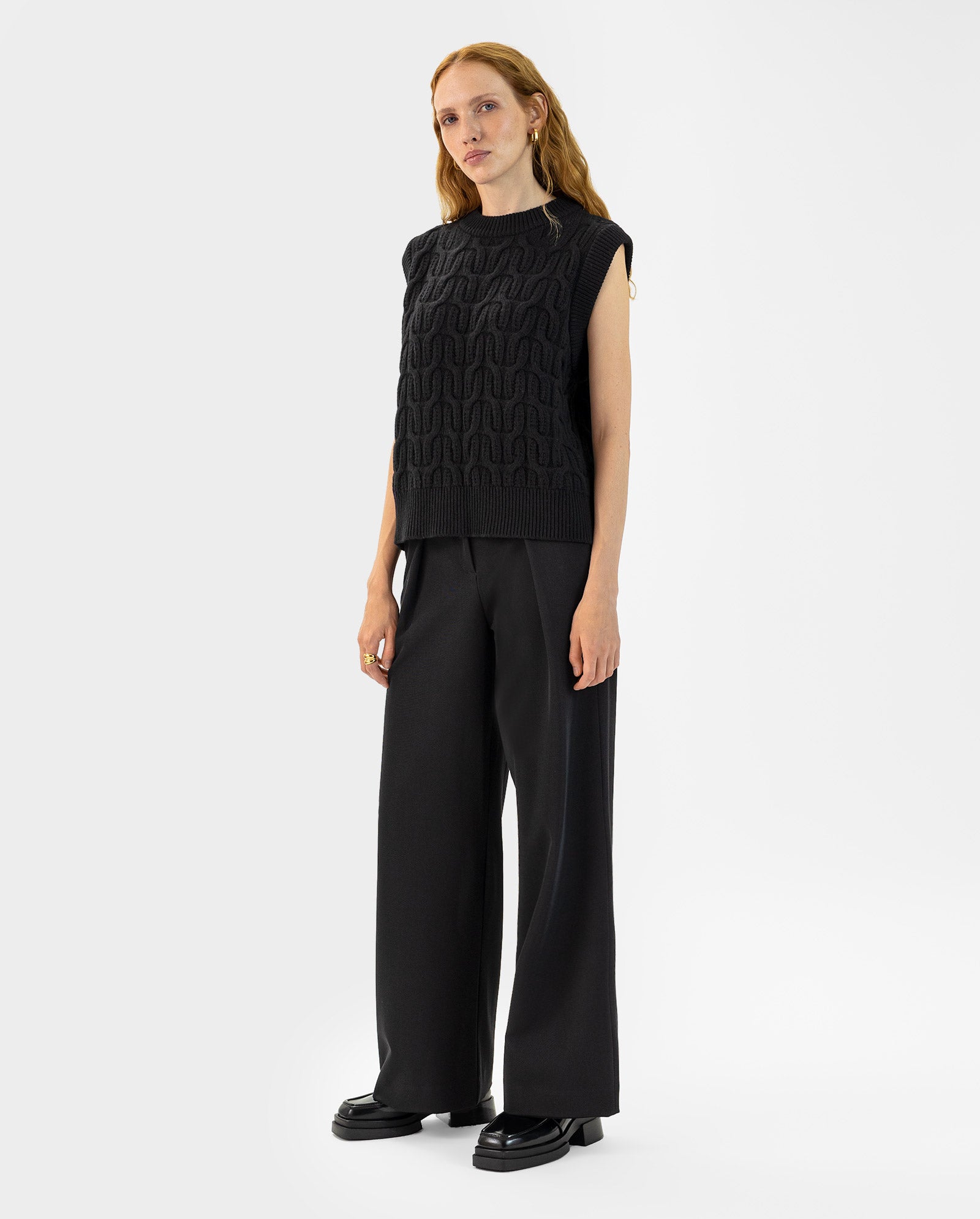 A woman stands against a white background wearing the IVY OAK KLARISSA Pullunder, a black sleeveless knit in extrafine merino wool, paired with wide-leg black trousers.
