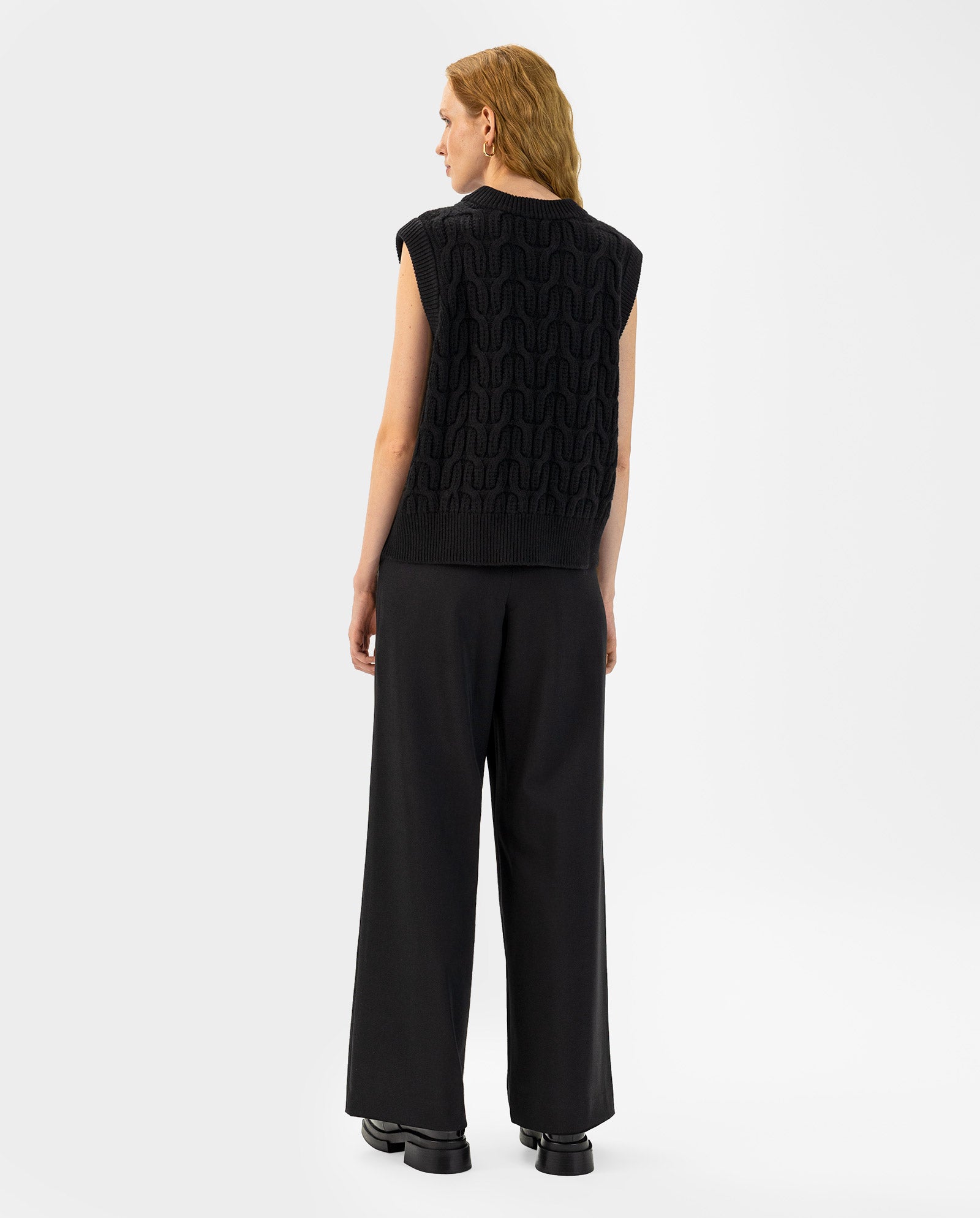 Person with wavy blonde hair, seen from behind, wears the IVY OAK KLARISSA Pullunder—a sleeveless black knit in extrafine merino wool—paired with wide-leg black pants.