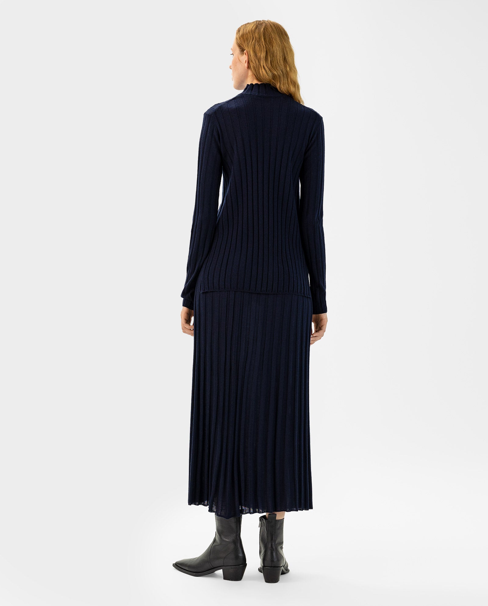 A woman stands facing away, wearing the IVY OAK KACY Jumper— a long-sleeved, navy Merino wool dress— paired with ankle boots.