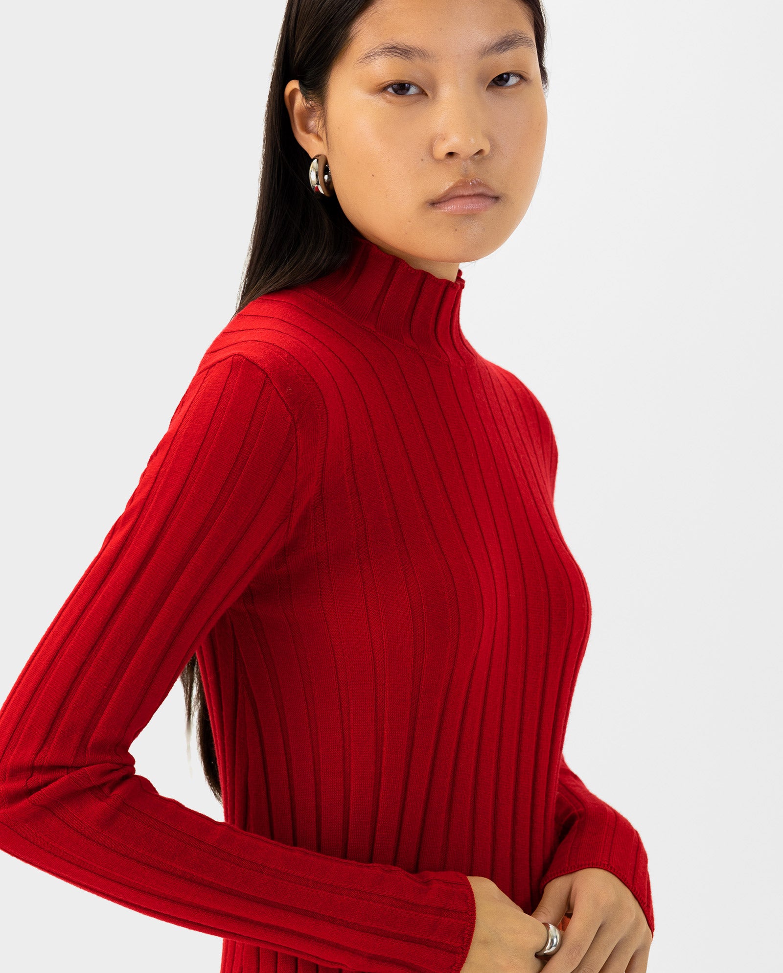 Woman with long dark hair wearing the red KACY Jumper by IVY OAK, featuring merino wool and a ribbed texture, paired with silver hoop earrings.