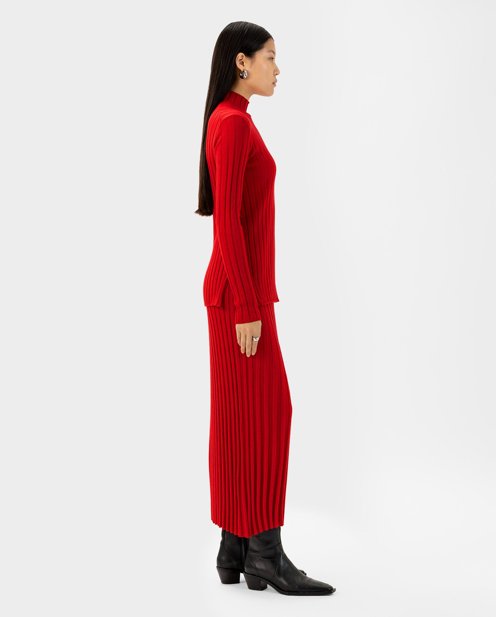 A woman in the IVY OAK KACY Jumper, a long-sleeve ribbed red dress with Rippstruktur, is paired with black boots and shown in profile against a white background.