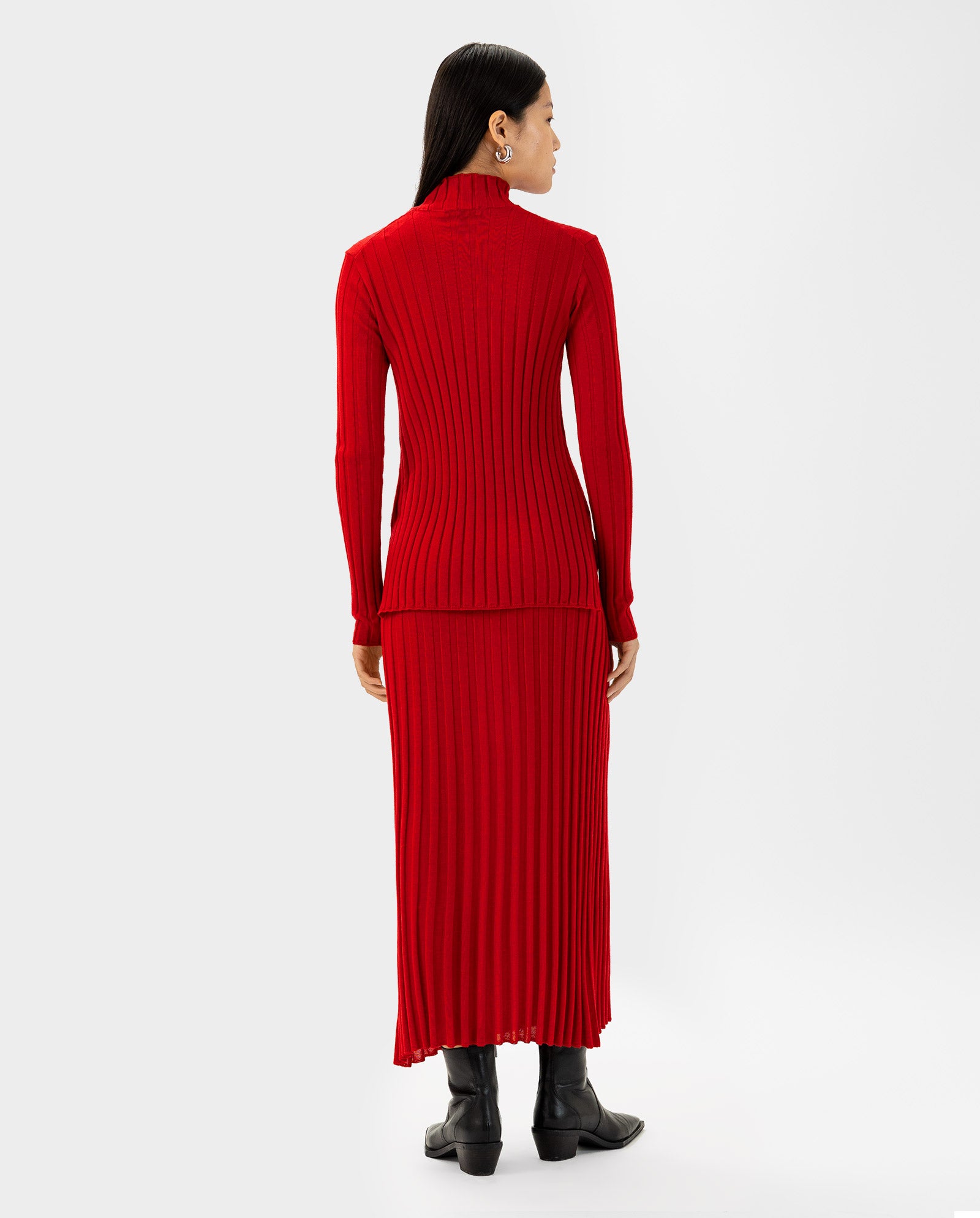 Woman wearing the IVY OAK KACY Jumper, a long-sleeved red dress with ribbed texture made from soft Merino wool, paired with black boots and standing with her back to the camera.