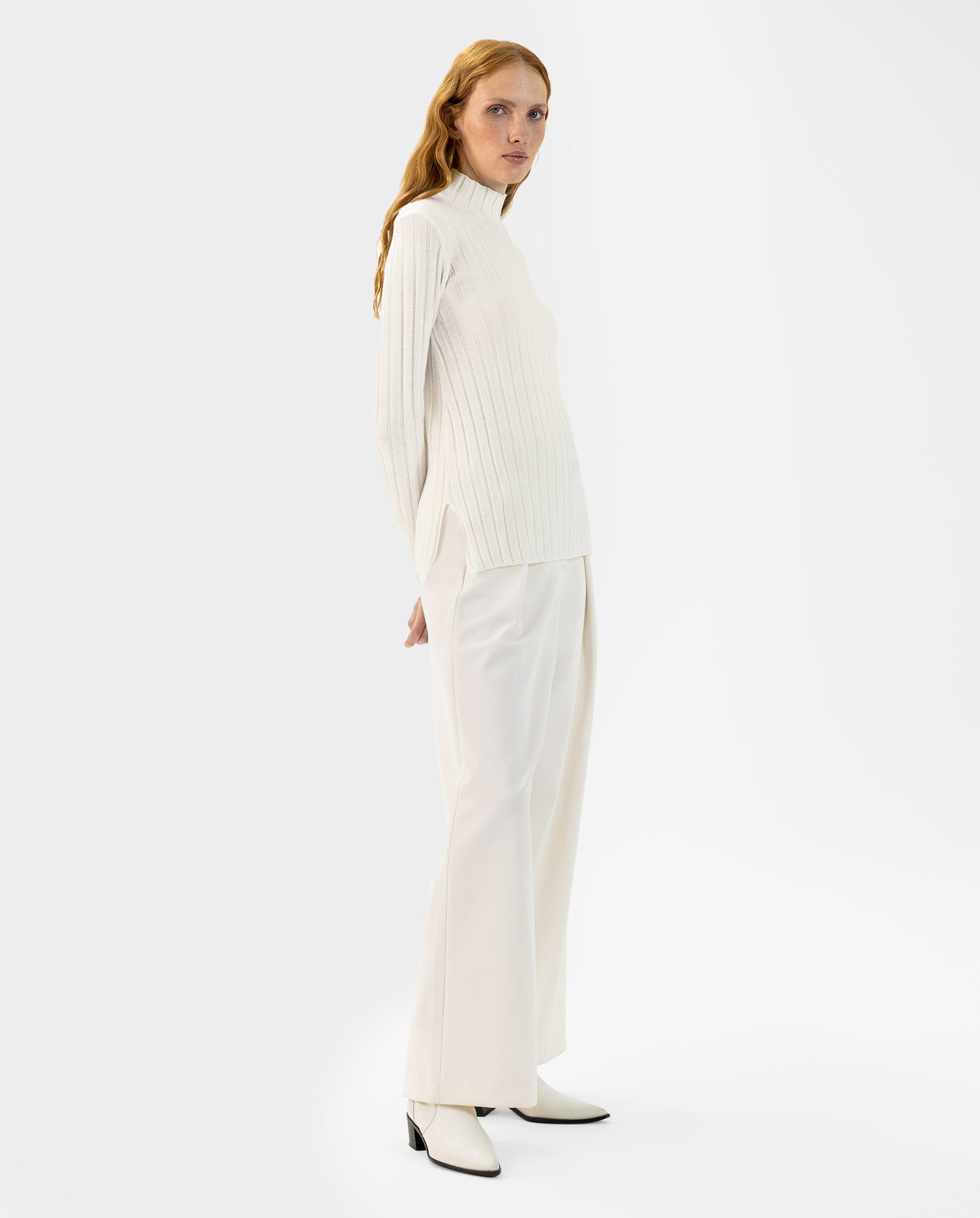 A woman wearing the IVY OAK KACY Jumper, white wide-leg pants, and white boots stands sideways.