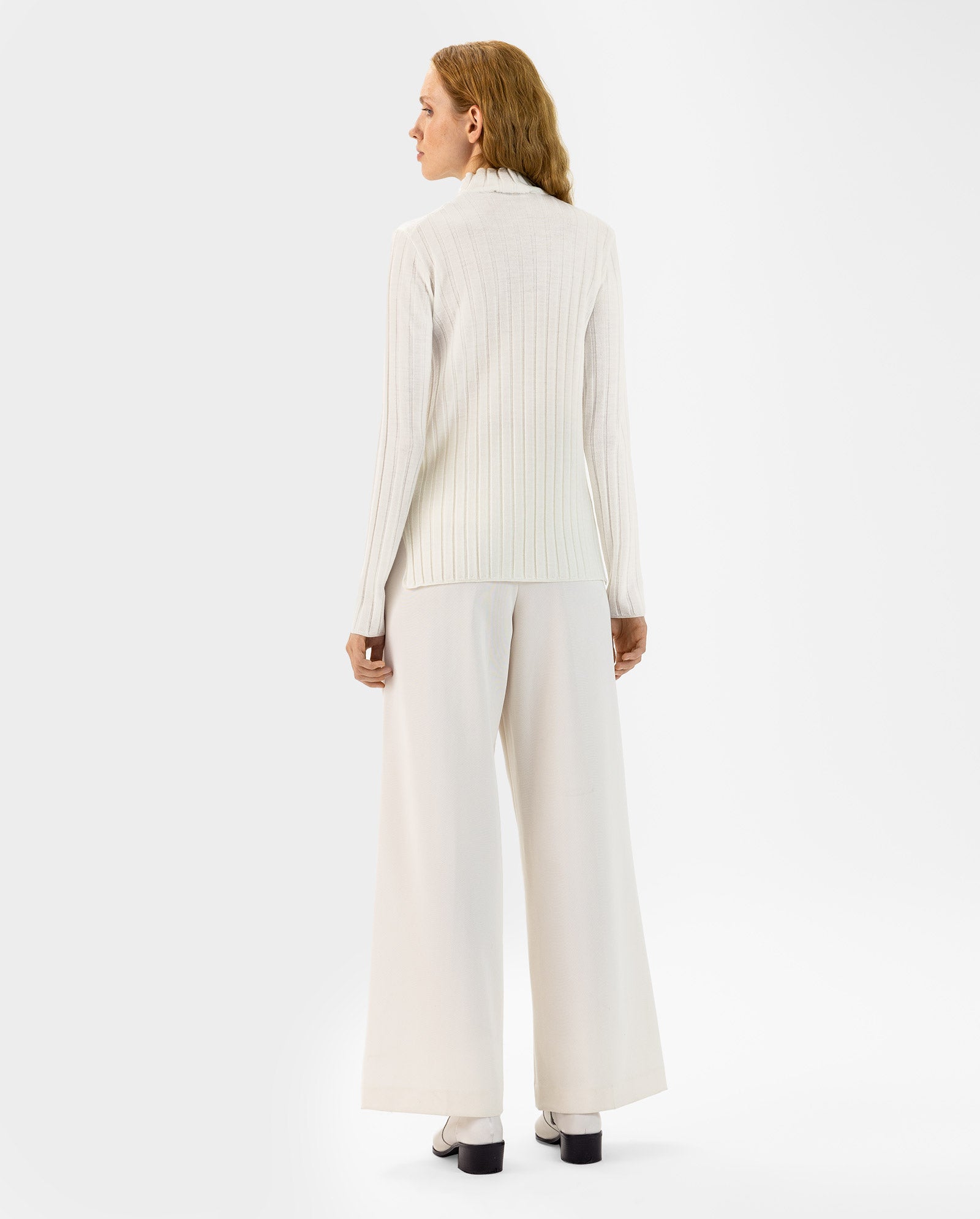 Woman with wavy hair, wearing the white KACY Jumper by IVY OAK and wide-leg pants, stands with her back to the camera.